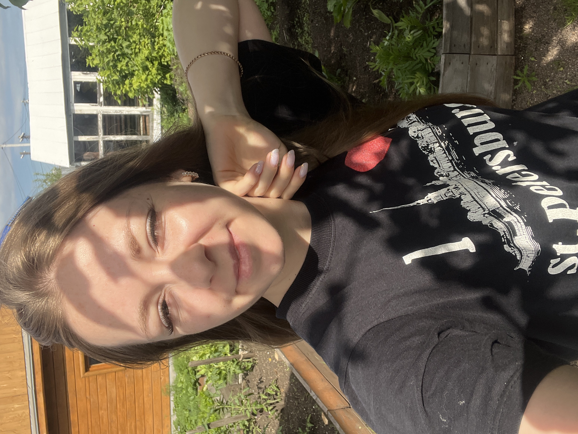 A young woman is pictured in a selfie taken outdoors. She has long brown hair and is wearing a black t-shirt with a graphic of St. Petersburg. She is in a garden setting, with raised garden beds, greenery, and wooden buildings visible in the background. The lighting suggests it is a sunny day. No specific landmarks or clues point to the city of Shadrinsk, Russia.