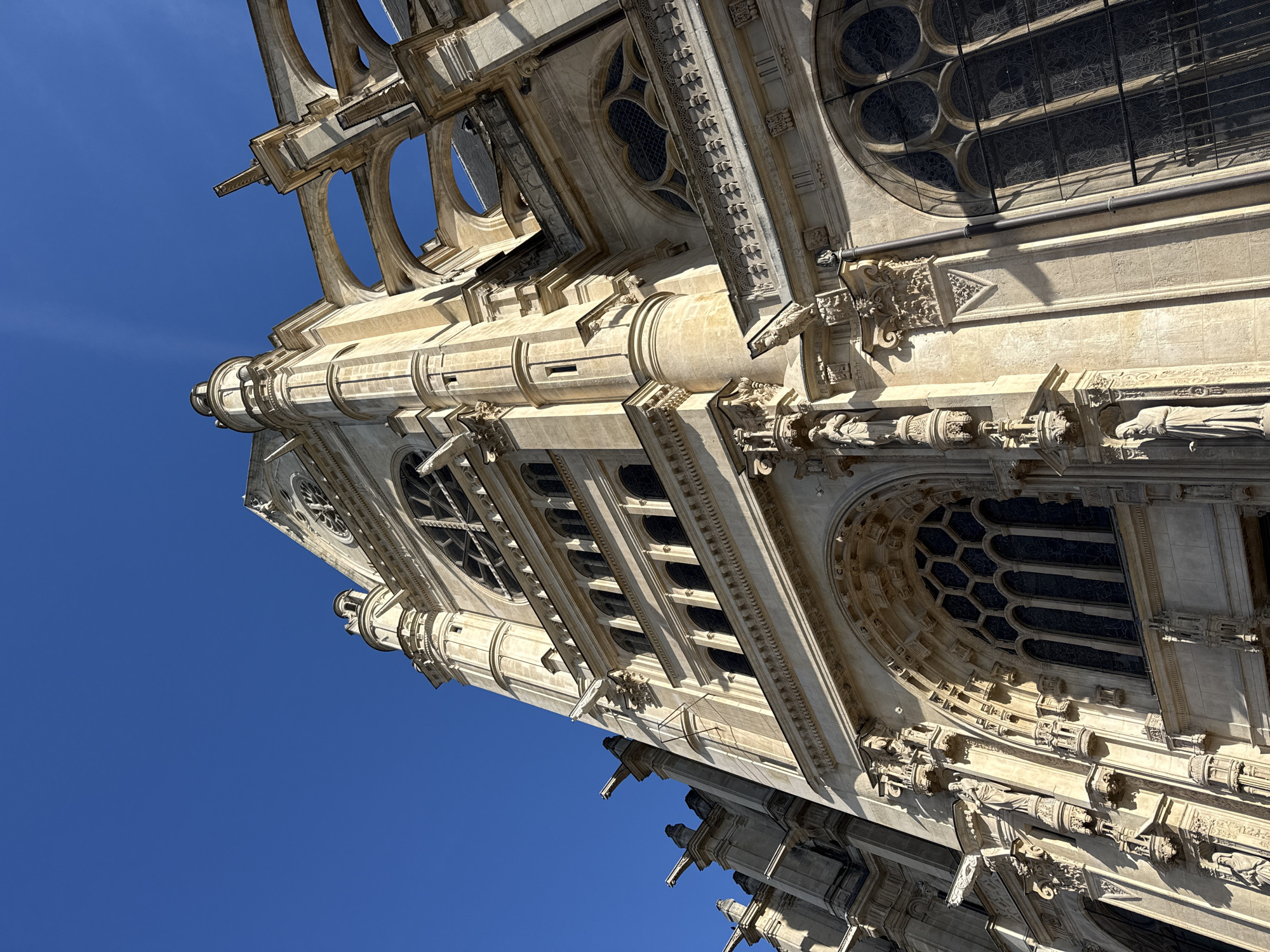 This is a low-angle view of a Gothic-style building, likely a cathedral, in Paris, France. The building is made of light-colored stone. The architectural details are elaborate, including pointed arches, circular windows with intricate tracery, ornate carvings, and gargoyles. The blue sky is visible in the background.