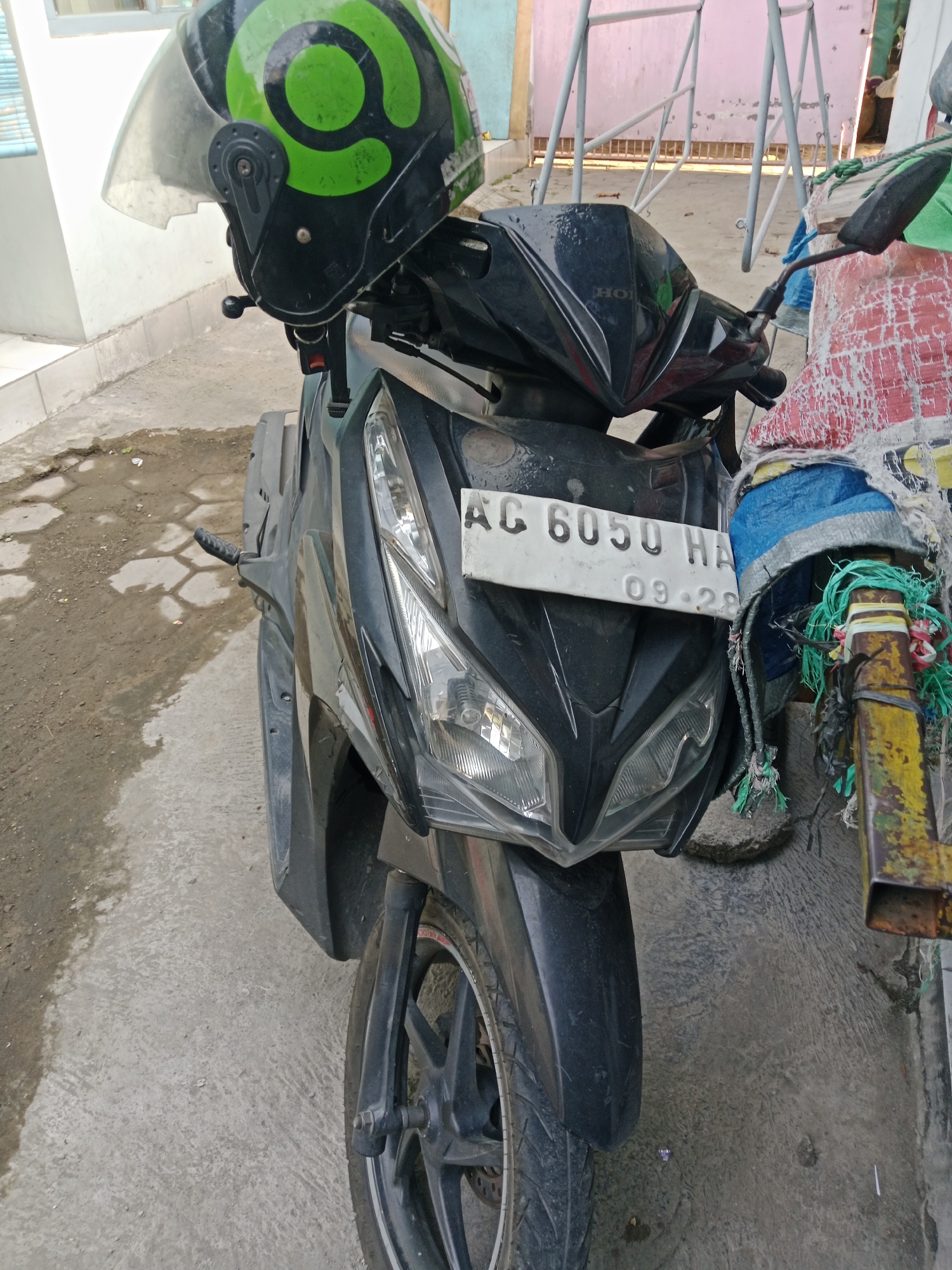 A black motorcycle with a green and black helmet on its handlebars is parked on a concrete surface. The license plate reads "AC 6050 HA" with "09-28" below it. A yellow metal post with various ropes and fabric tied to it stands to the right of the motorcycle. In the background, a light-colored building and a metal gate are visible. The ground shows patches of dirt and debris. The overall scene appears to be an outdoor area in a residential or commercial setting.