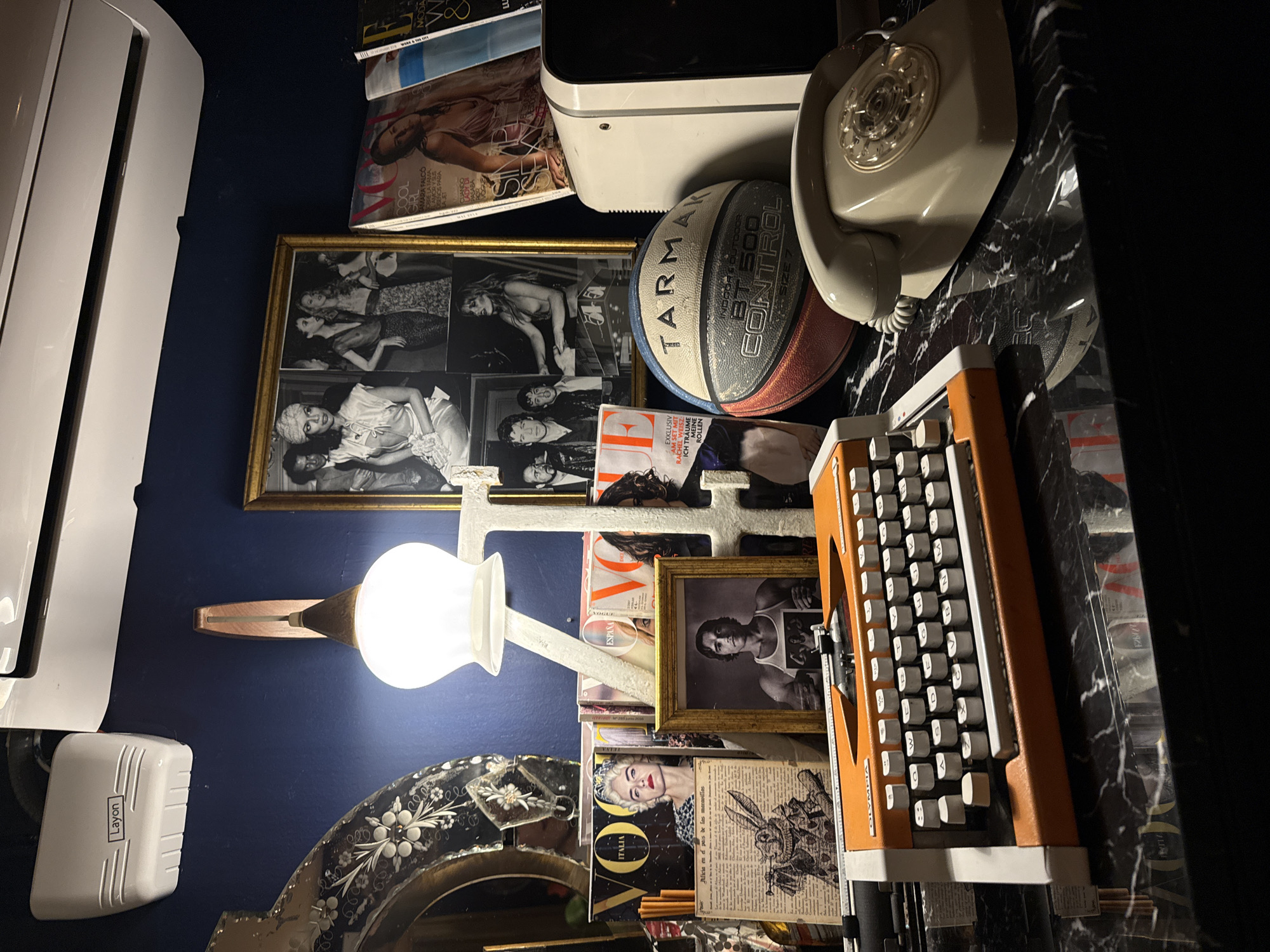 The image shows a cluttered interior setting. On the left, there is a white air conditioning unit labelled "Layon" mounted on a dark blue wall. Next to it is a wall-mounted lamp with a white shade, shining on a collection of items. There are framed photographs, including black-and-white portraits, stacked together. Assorted magazines, including issues of "Vogue Italia" and "Vogue España," are scattered among the framed photos. On top of the stack are a beige rotary telephone and a basketball. The right side features a vintage orange typewriter placed on a dark marble surface. The environment is dimly lit.