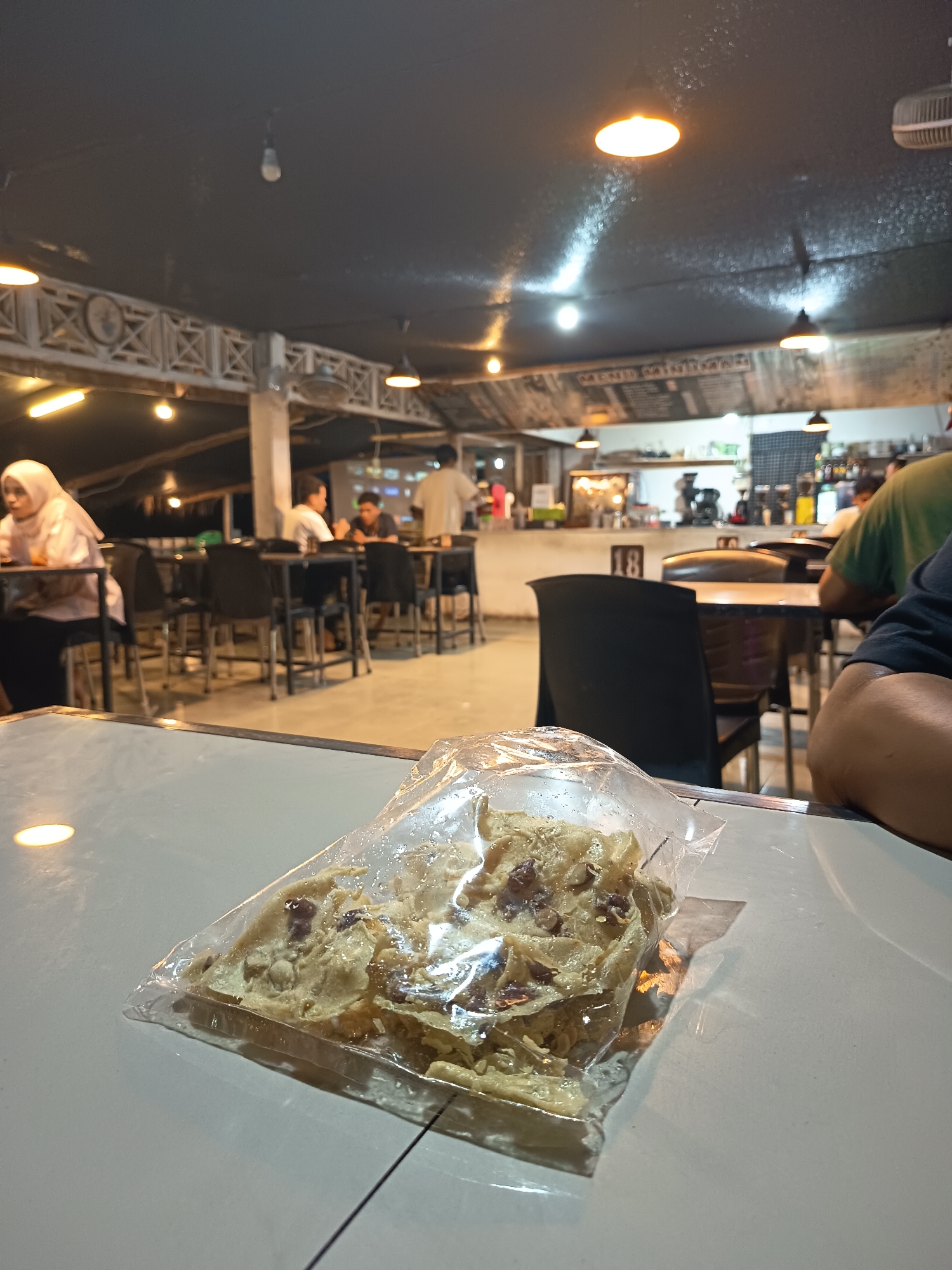 This media file depicts an indoor scene within a cafe or restaurant in Singkil, Indonesia. The setting appears to be evening, indicated by the artificial lighting from overhead lamps that cast a warm glow. The ceiling is dark, with several lights visible, some emitting a warm, yellowish light and others cooler white. Decorative, latticed panels are visible along the upper part of the wall, adding an architectural detail to the space.
In the foreground, on a white table, is a clear plastic bag containing a snack. The snack appears to be a type of crispy cracker or wafer, light in color and possibly containing small, dark pieces. The bag is slightly crinkled and reflects the ambient light.
The mid-ground shows several tables and chairs, some occupied by patrons. To the left, a person wearing a light-colored hijab is seated at a table. Further back, other individuals are visible, some engaged in conversation. The tables are dark, and the chairs are black plastic.
The background reveals more of the cafe's interior, including what looks like a counter or serving area with shelves stocked with items. There are also more tables and chairs, suggesting a moderately busy environment. The overall mood appears relaxed and casual, typical of an evening gathering at a local eatery. There is no visible text that can be deciphered in the image.