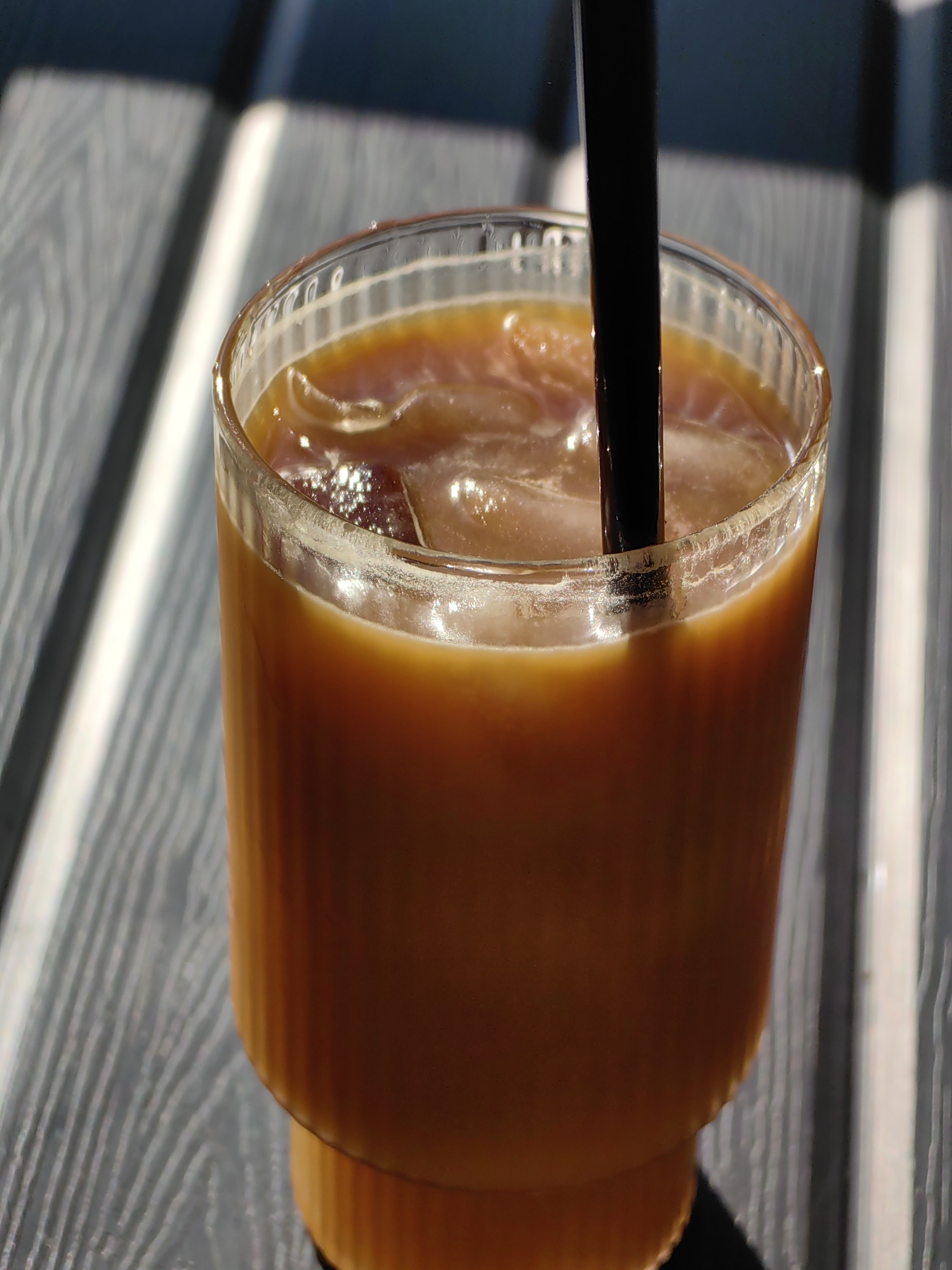A close-up shot depicts a clear glass filled with an iced coffee beverage, complete with a black straw, sitting on a table with grey wooden slats. The setting is outside under bright sunlight, creating distinct shadows across the table. The image suggests an outdoor café or patio environment. The beverage appears to be cooling, with some condensation visible on the glass.