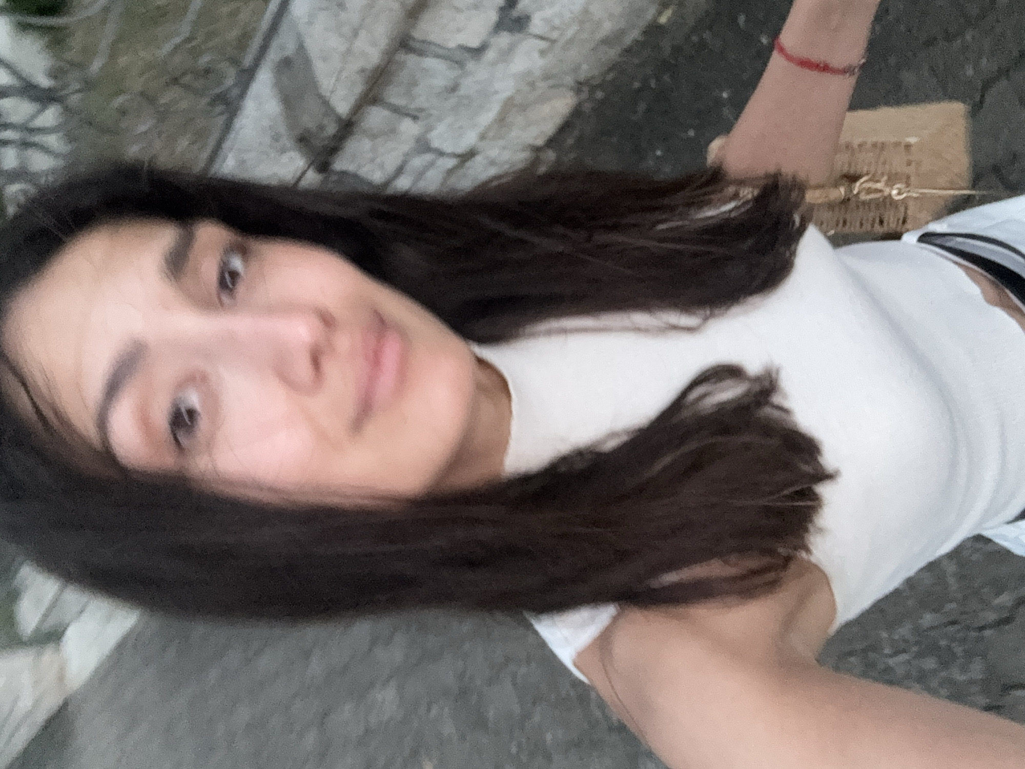 The image is a selfie taken by a person with dark hair, wearing a white top. The person's face is partially visible, with their right eye looking towards the camera. They are also wearing a thin red bracelet on their left wrist. A woven bag is visible behind their arm. The background appears to be an outdoor setting, with a textured surface that could be pavement or stone. The lighting suggests it might be daytime, but the overall quality of the image is somewhat blurry and overexposed. The scene is framed from a high angle, looking down at the subject. There is no visible text in the image. The context of Bar, Montenegro is provided, but there are no specific visual cues in the image to confirm this location.