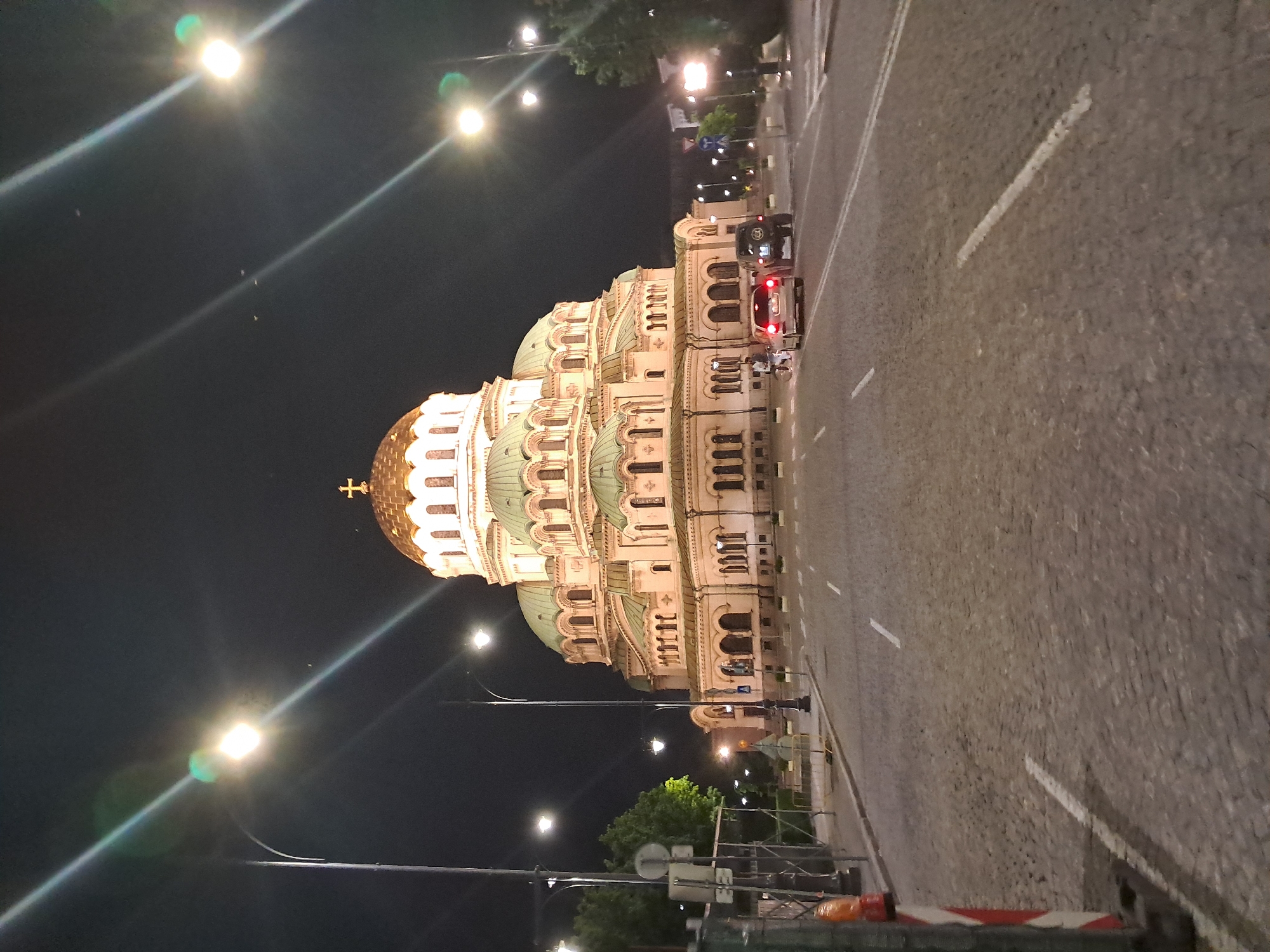The image captures the Alexander Nevsky Cathedral in Sofia, Bulgaria at night. The cathedral is brightly lit, showcasing its architectural details. A street with cars passes alongside the cathedral. The pavement is visible in the foreground. Streetlights line the road, adding to the illumination. The sky is dark, emphasizing the illuminated cathedral and street scene.
