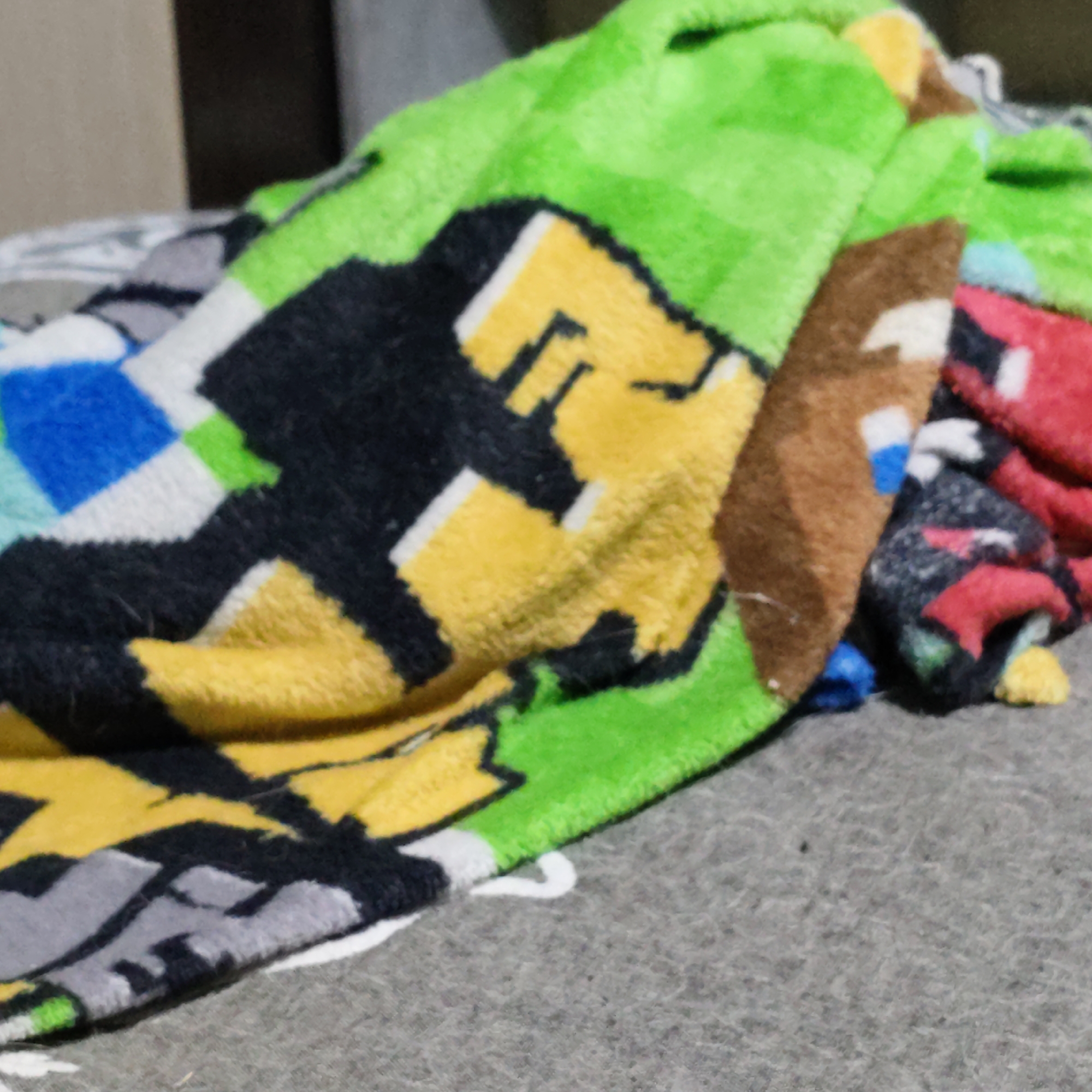 A close-up, blurry, high-angle shot shows a pile of folded, plush blankets with a pixelated, video game-inspired pattern. The dominant colors are bright green, yellow, black, white, brown, and red. The blankets are layered on top of a gray, textured surface, possibly a carpet or another blanket. The lighting is dim, casting soft shadows. The composition is tight, focusing on the textures and patterns of the fabric. The background is out of focus, suggesting a domestic setting.
