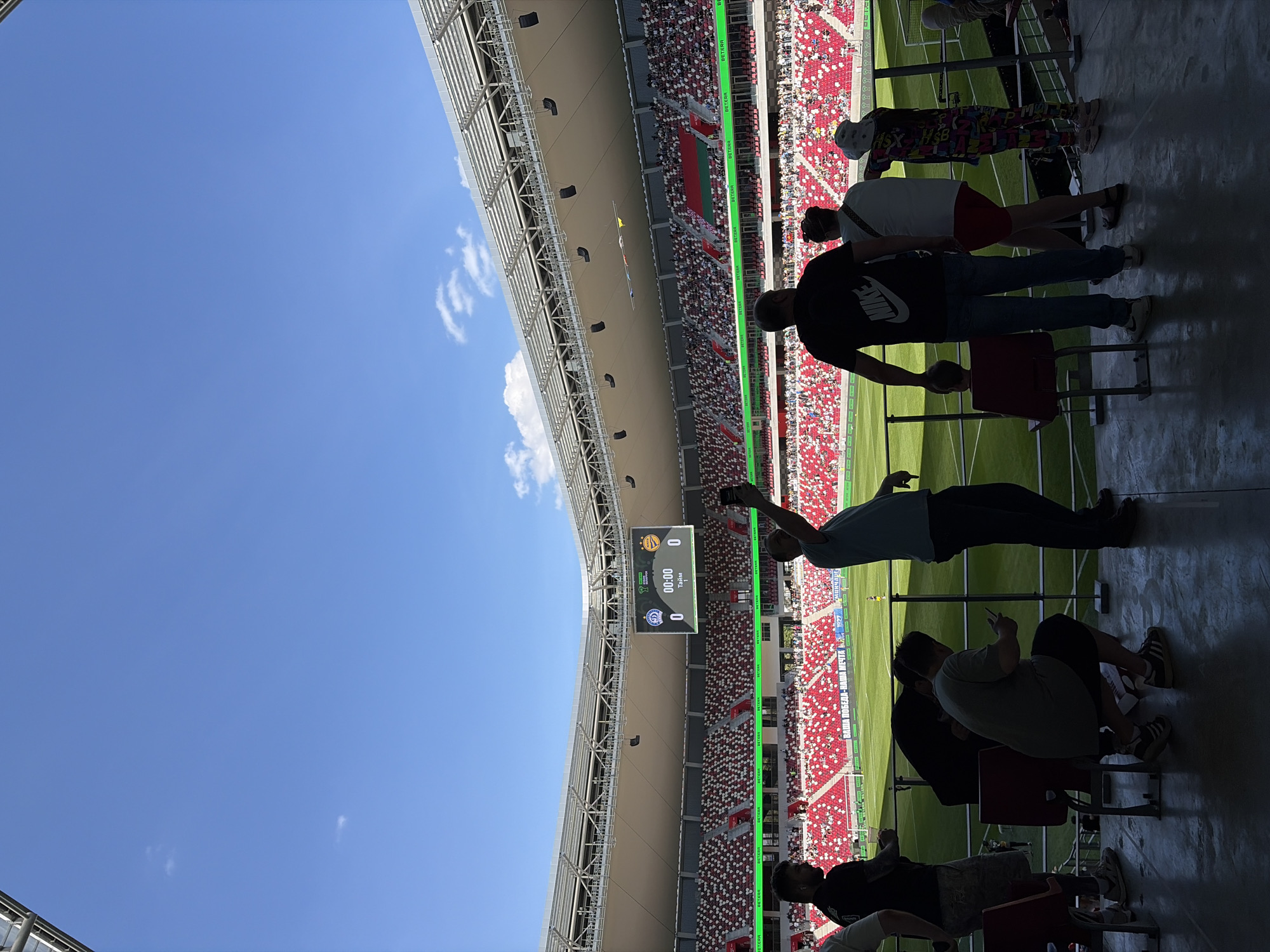 The image shows a view from a stadium, possibly in Minsk, Belarus, as the scoreboard shows Russian words and the match is at 0-0. There are spectators in the stands, and some are standing near the camera taking pictures. The sky is blue, and the weather appears sunny. The stadium is mostly full.