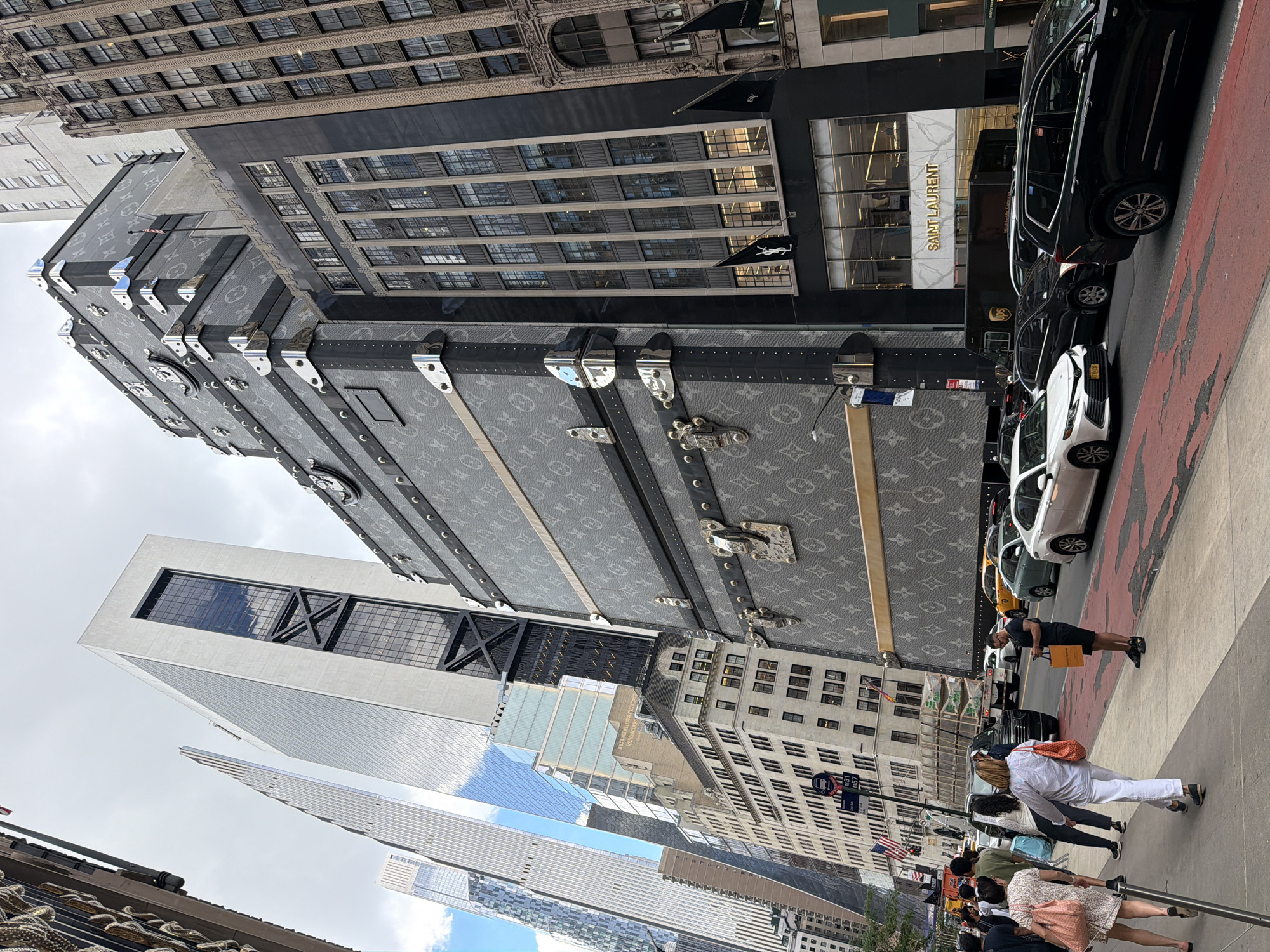 This image captures a street scene in New York City, likely Midtown Manhattan, given the iconic architecture and presence of high-end retail. The focal point is a colossal Louis Vuitton trunk facade, part of a building that also displays a "SAINT LAURENT" sign. The trunk is elaborately decorated with the brand's signature monogram and chrome accents, towering over the street.
Several modern and classic buildings rise in the background, reflecting the city's diverse architectural landscape. These include sleek glass skyscrapers and older, ornate buildings. The street itself is bustling with activity. Cars, including a white Toyota Camry and a black SUV, are on the road, some of which are yellow taxis, common in New York City. Pedestrians are walking on the sidewalk. One person is holding an orange shopping bag, suggesting recent purchases. Another individual is looking at their phone.
The sky is overcast, indicating a cloudy day. The sidewalk features a red-striped design, possibly indicating a bus lane or a pedestrian zone. The overall impression is a vibrant urban environment, characteristic of New York City's commercial districts, where luxury brands are prominently displayed.