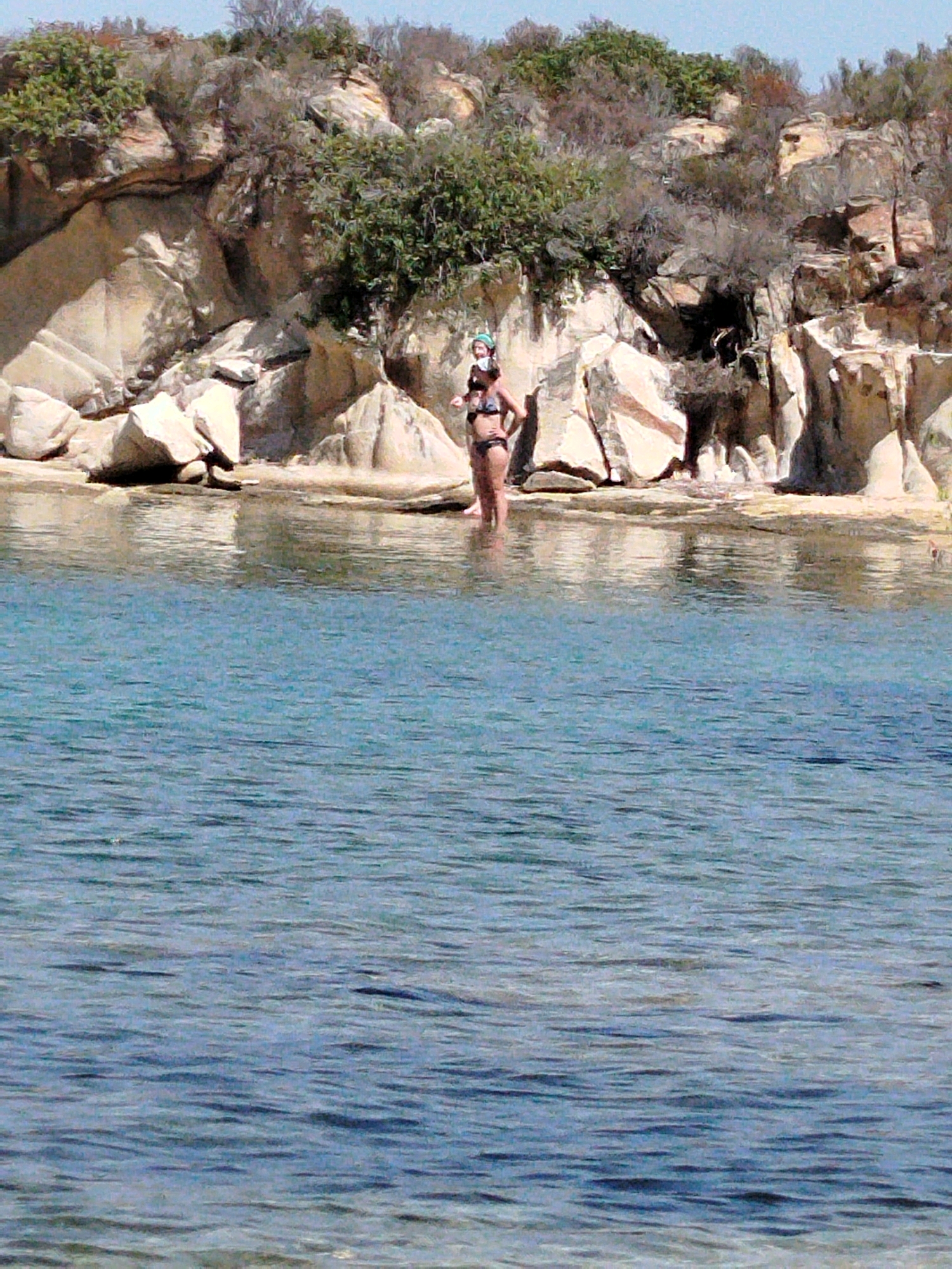 A woman stands in shallow, blue water, wearing a dark bikini and a snorkeling mask. She is positioned in front of a rocky shoreline with sparse vegetation under a clear sky. The water is calm, with visible ripples and reflections of the rocks. The background consists of large, light-colored rock formations and dry brush. The scene appears to be a natural, coastal environment, likely in Sithonia, Greece, a region known for its scenic beaches and rocky shores.
