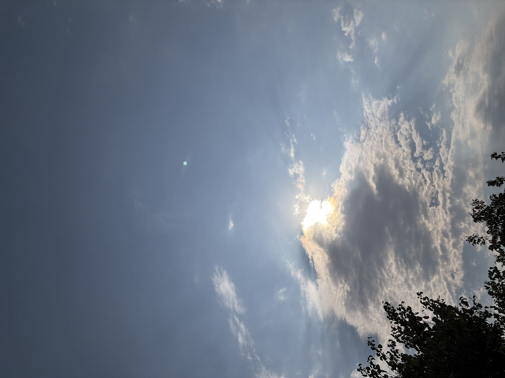 The image shows a sky with scattered clouds and the sun peeking through. A faint green circular light artifact is visible in the upper right quadrant of the sky. The lower portion of the image is occupied by the silhouettes of trees. The location is specified as Oxentea, Moldova.