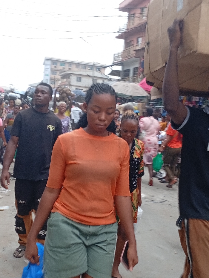 A crowded market scene shows numerous people walking amidst stalls. A young woman in an orange shirt and green shorts walks in the foreground, looking down. To her left, a man in a black shirt walks beside her. Behind them, people are carrying items on their heads and shopping. A building is visible in the background. The ground is littered with debris. Another man carrying a large box passes by on the right.