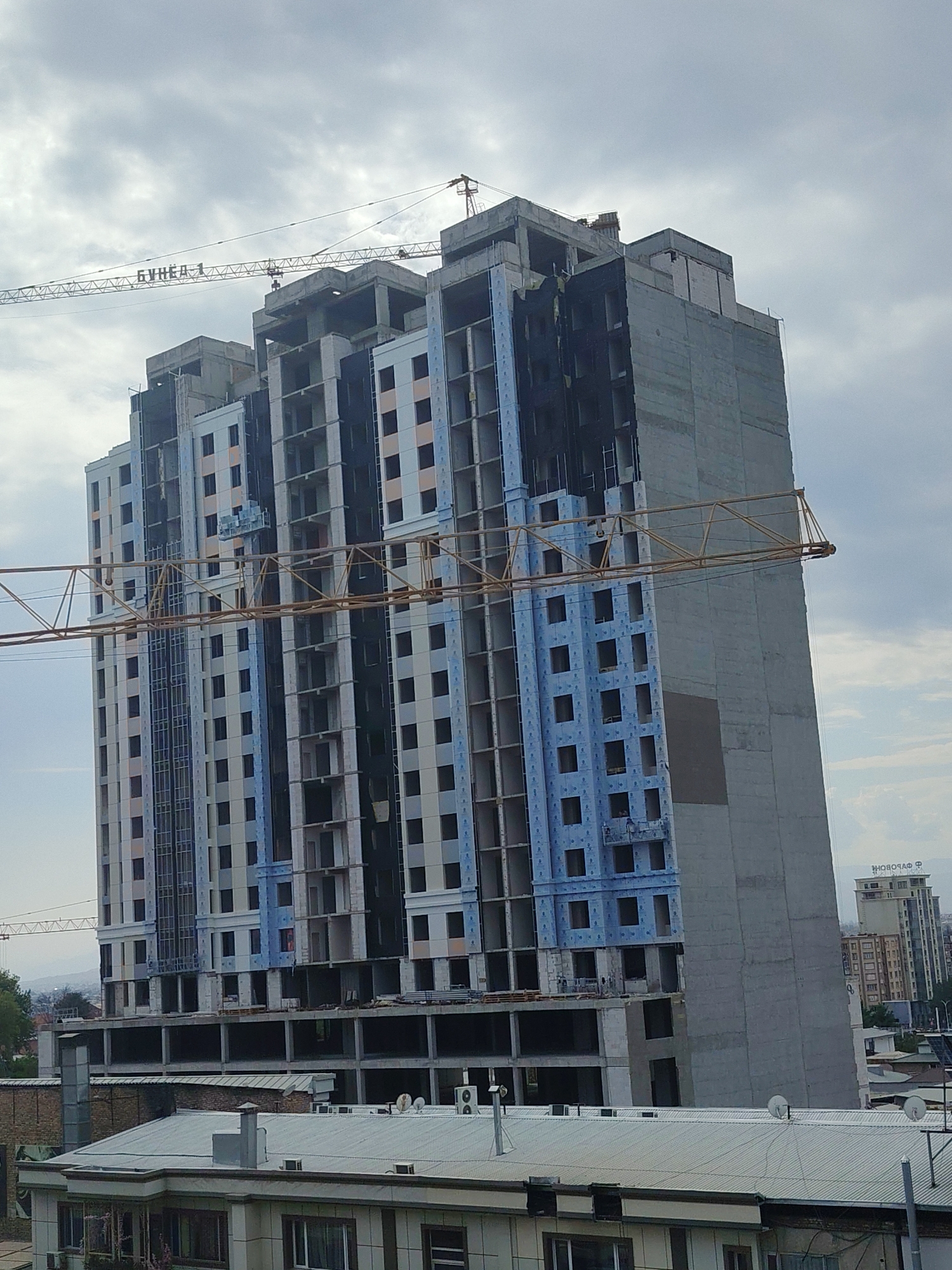 A construction crane is visible with the Cyrillic text "БУНЕА 1" on its jib. A tall, unfinished building, likely an apartment complex, dominates the foreground. The building's facade shows rows of dark rectangular window openings and some sections covered in blue insulation material. A construction elevator or gondola is attached to the side of the building. In the background, other buildings and a distant cityscape are visible, suggesting an urban environment. The sky is overcast with clouds. The scene appears to be the city of Khujand, Tajikistan, indicated by the presence of Cyrillic text and architectural styles typical of the region.