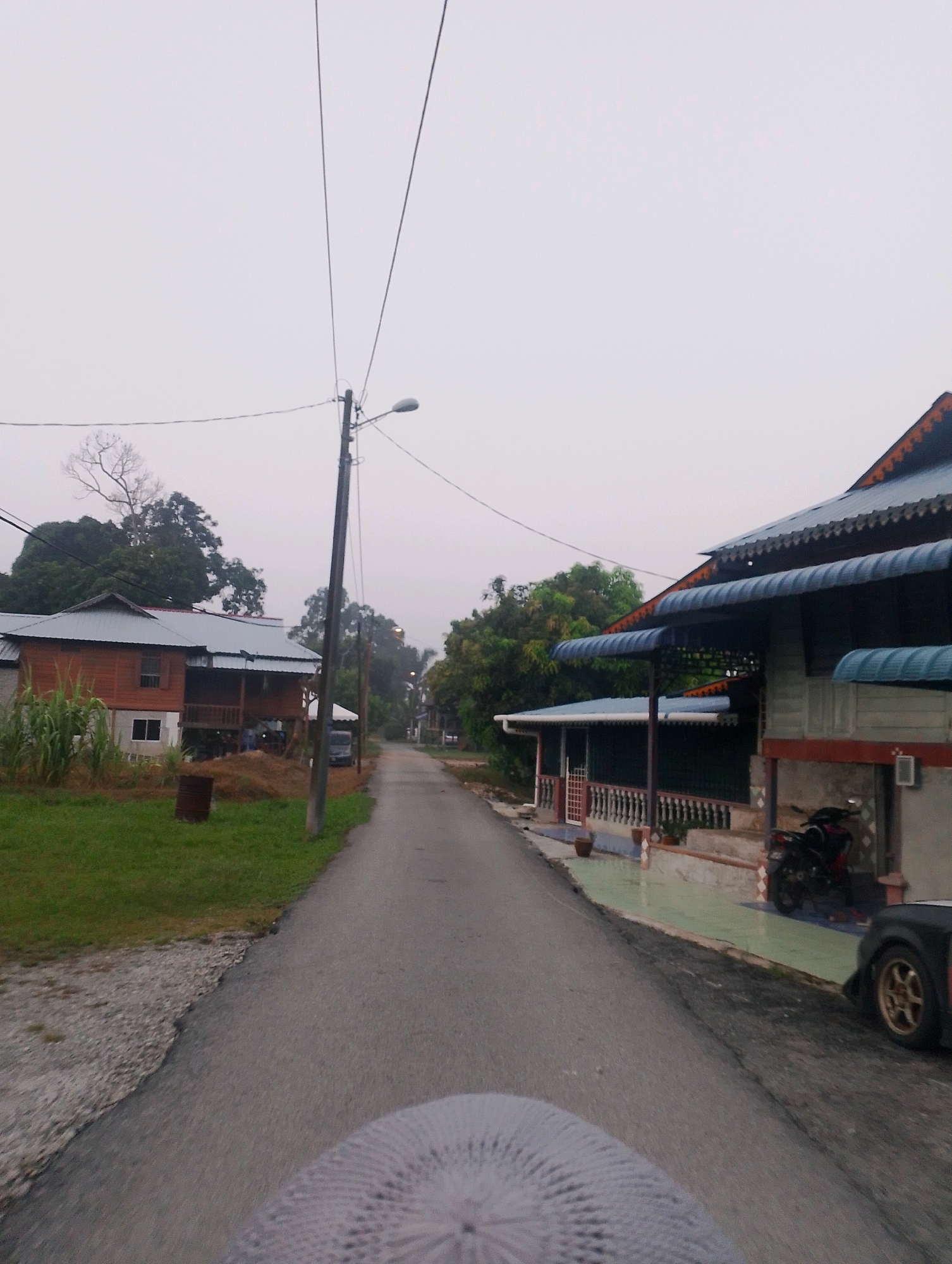 The image depicts a view from a moving vehicle, likely a motorcycle given the angle and the presence of a textured object in the foreground that appears to be part of a seat or cover. The scene is a rural road in Pulau Tiga, Malaysia. On the left side of the road, a wooden house on stilts is visible, with green grass and tall plants in front. To the right, another house with a multi-tiered, dark blue awning is situated. A motorcycle is parked in front of this house. Further down the road, more houses and trees can be seen, with power lines stretching across the overcast sky. The road itself is paved and appears to be in fair condition. The overall atmosphere suggests a quiet, potentially early morning or late afternoon setting due to the subdued lighting and the cloudy sky.