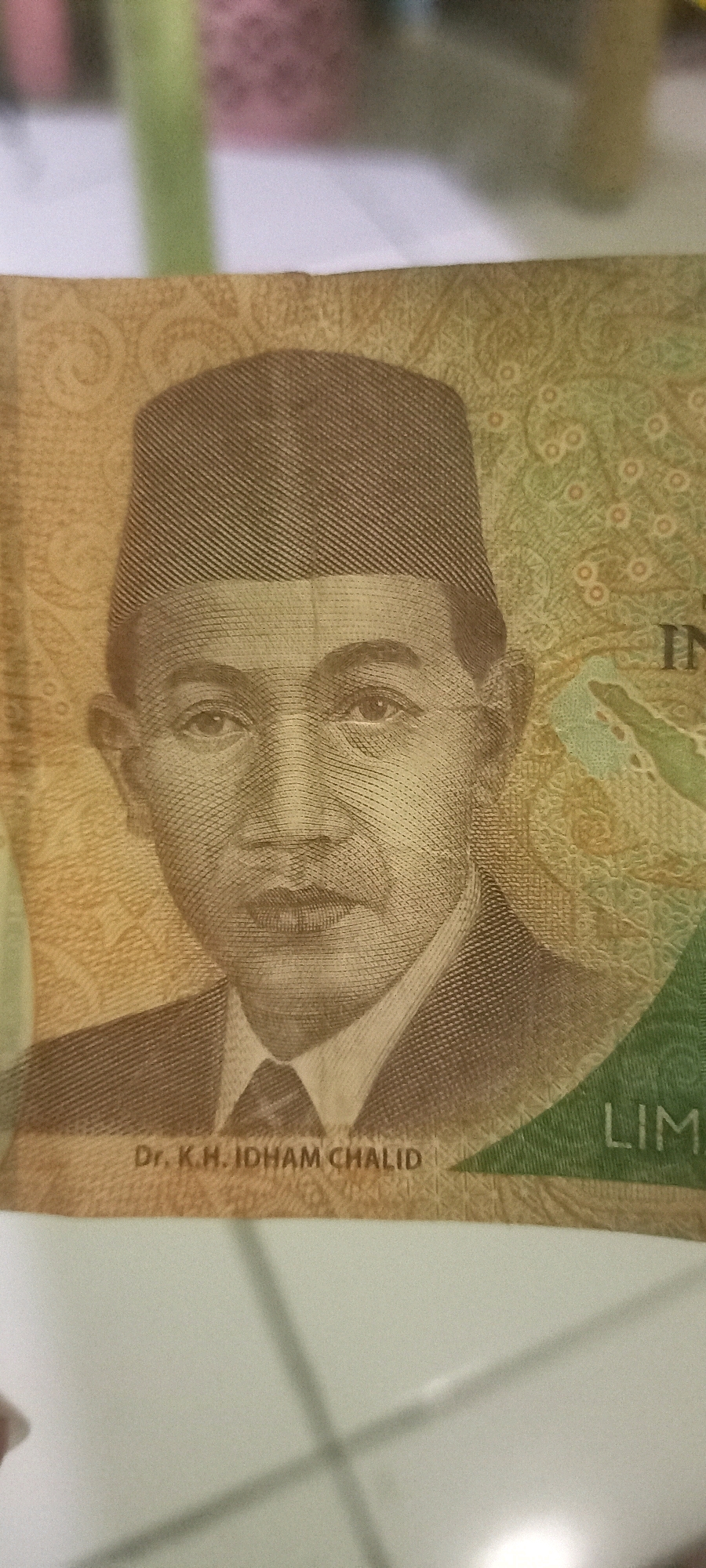 The image displays a close-up of an Indonesian banknote. The prominent feature is a portrait of a man wearing a dark, patterned cap and a suit. His name, "Dr. K.H. IDHAM CHALID", is printed below his portrait. The banknote has a textured pattern in shades of yellow and green, with the word "LIM" visible on the right side. The background is blurred, showing hints of a light-colored surface. There is no discernible indication of the city of Argasoka, Indonesia, within the image itself.