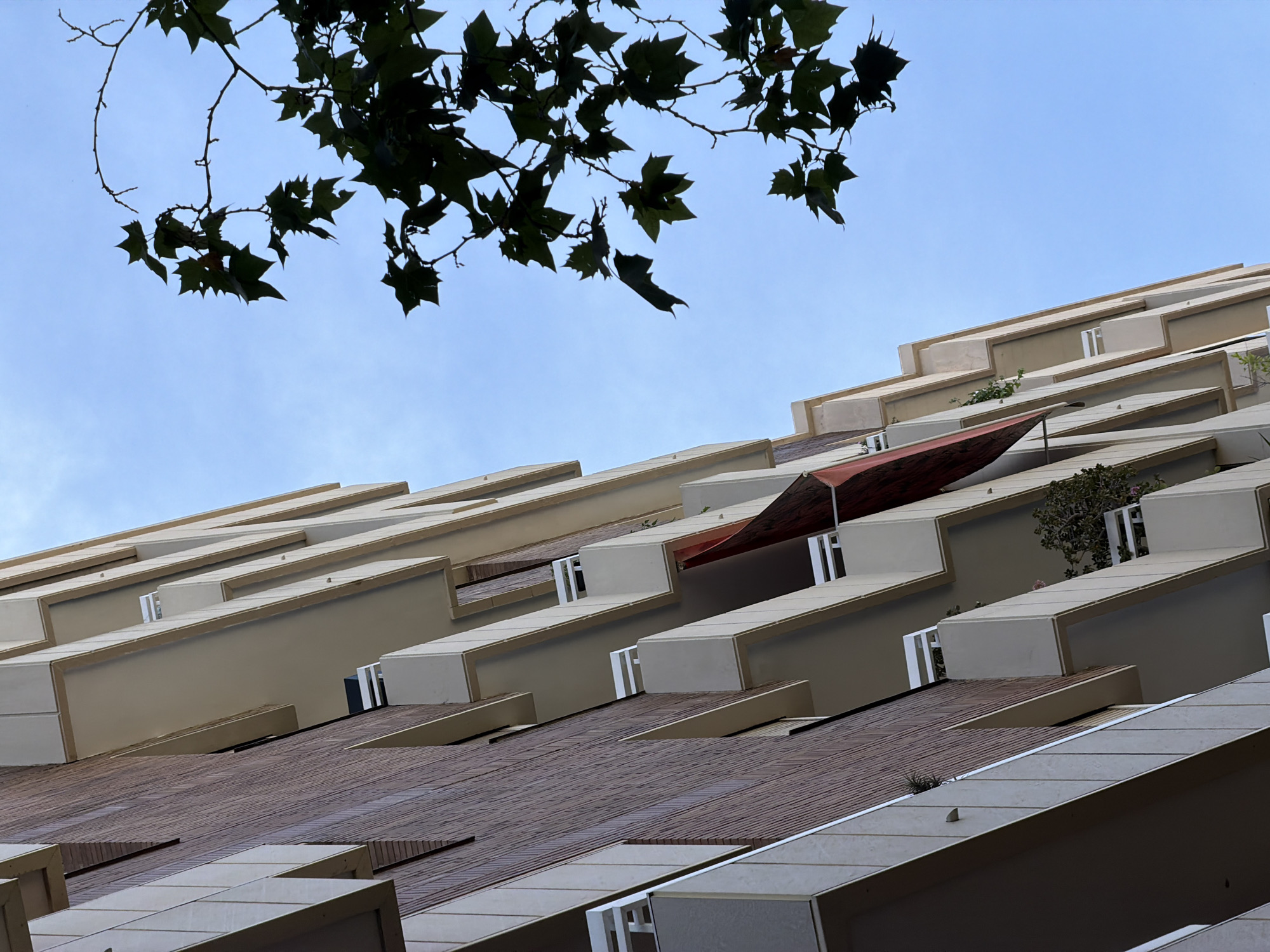This image captures a low-angle view of a modern apartment building with a brick facade and cream-colored balconies against a clear blue sky. A tree with dark green leaves is silhouetted against the sky in the upper right portion of the frame. The building features multiple levels of balconies, each with white railing. A red awning or shade is partially visible on one of the balconies. There are some plants on the balconies. The architecture suggests a contemporary urban setting. This building is located at 7-9 Carrer de la Selva de Mar in Barcelona, Spain.