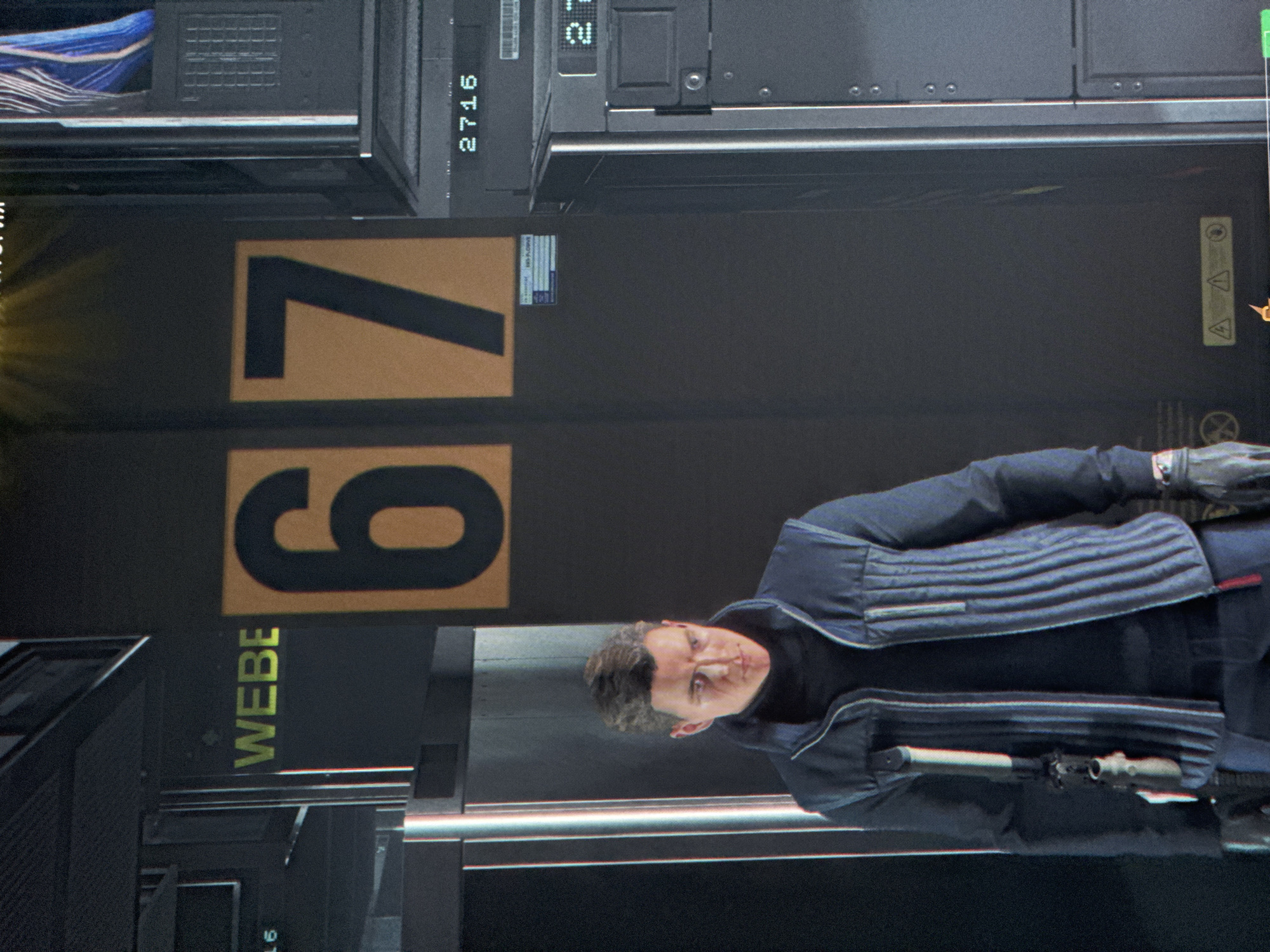 The image shows a man in a dark blue jacket and black turtleneck standing in front of a large, dark panel with two large orange numbers, "7" and "9", displayed vertically. To the left of the numbers is a vertical yellow text that reads "WEBE". Above the "7" and to the left, some blue and white striped material is visible. To the right of the "7", a small label with text is visible. Above the "7", some black metal structures with circular indentations are visible. A digital display showing "2716" is mounted vertically on a black surface above the "7". There is a warning symbol with an exclamation mark inside a triangle visible on the dark panel to the right of the "7". The man is looking towards the camera with a neutral expression. He has short, dark, styled hair and appears to be middle-aged. He is wearing gloves. In the lower right of the image, a portion of the man's dark jacket is visible, revealing a metallic object protruding from his hip. The overall scene appears to be industrial or futuristic, possibly a set from a science fiction film or video game, with a dark and utilitarian aesthetic. The lighting is somewhat dramatic, highlighting the man and the large numbers. The provided location context is Moscow, Russia, but there are no direct visual cues in the image to confirm this. The image appears to be taken indoors, with artificial lighting.
