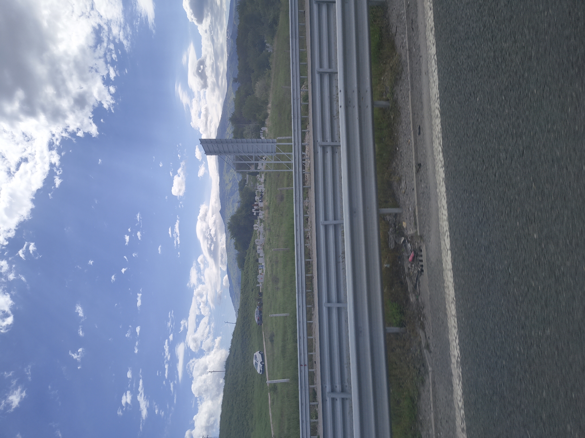 The image shows a view from a vehicle on a road near Kralev Dol, Bulgaria. To the left of the frame is a blue sky with scattered white clouds. In the distance are mountains. Adjacent to the road is a grassy field with what appears to be vehicles parked in it. A guardrail separates the road from the field. The road itself is asphalt and occupies a significant portion of the frame.