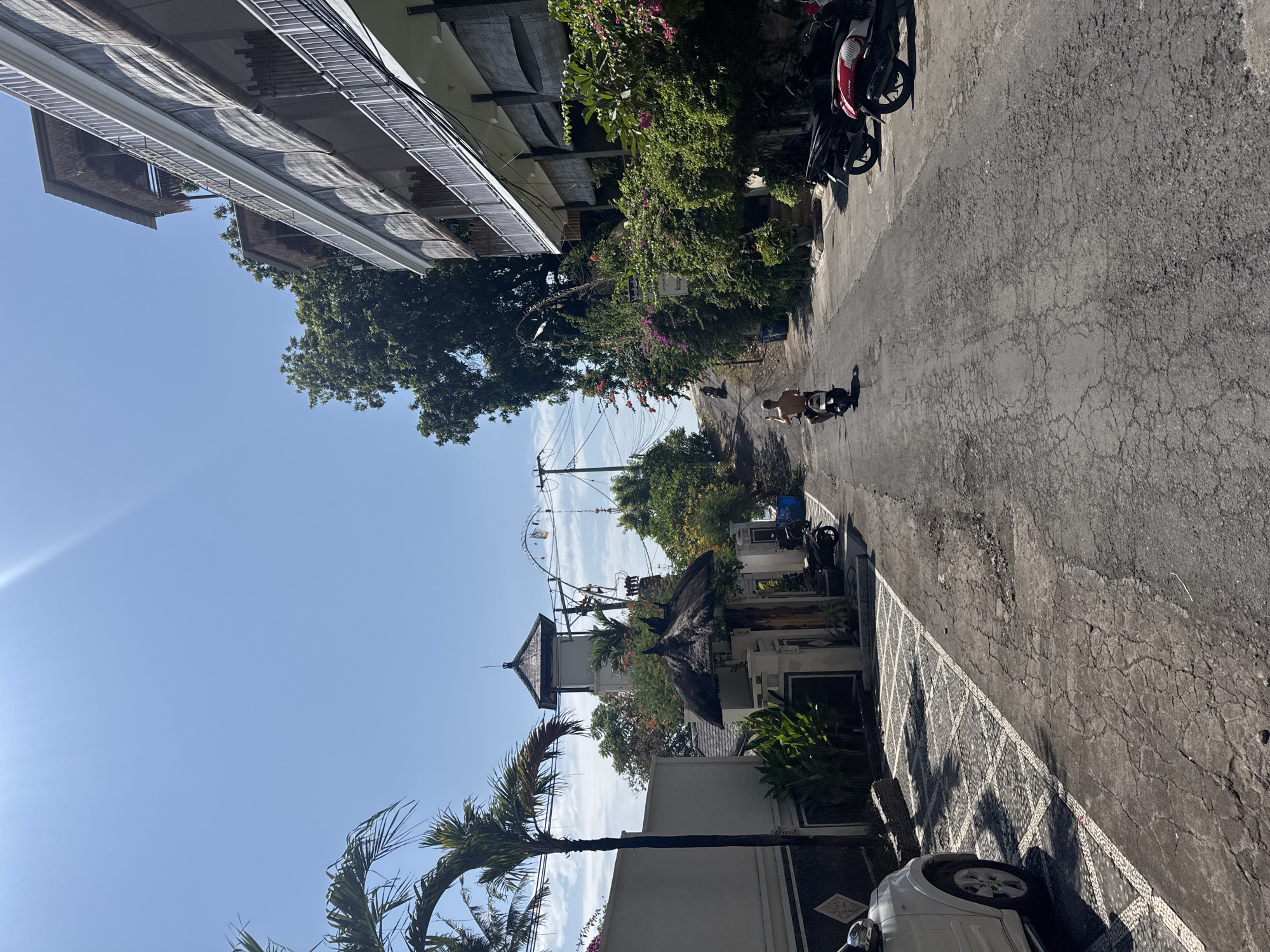 The image shows a narrow, cracked asphalt road in a tropical environment, likely a village or resort area, with buildings and lush vegetation on both sides. The road slopes upwards into the distance. On the left, a white SUV is parked, partially visible, next to a patterned paved area. Further up the left side of the road is a white wall and a tall palm tree, followed by a building with a traditional thatched roof entrance and a tall, square tower structure behind it. Electrical poles and wires are visible in the background. On the right side of the road, a multi-story building with balconies and woven bamboo-like coverings extends upwards, interspersed with dense green foliage and pink and purple flowering plants. Several motorbikes are parked on the right side of the road. A person is riding a motorbike up the road, seen from behind, wearing shorts. The sky is clear blue with a bright sun casting a glare. The general impression is a sunny day in a warm climate. The location is identified as Abang, Indonesia.