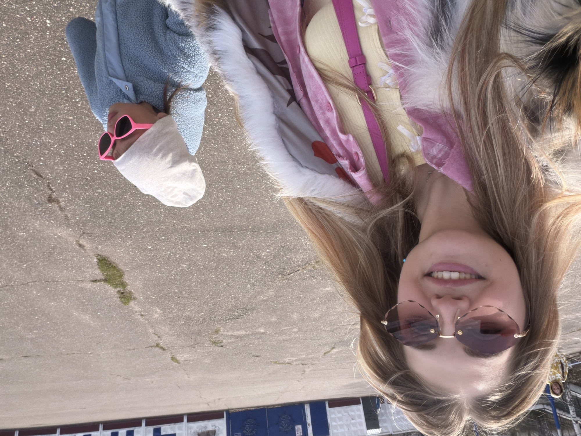 Two individuals are captured in an outdoor setting. The taller figure, a young woman, is smiling at the camera and is wearing sunglasses. Her long, brown hair flows beside her face. She is dressed in a yellow top under a pink button-down shirt with a fur-lined coat over both.
The second figure, a child, is positioned at the opposite edge of the frame. They are wearing pink sunglasses, a white hat, and a blue textured jacket.
The scene is set on a paved surface, likely a road or parking area. In the background, there is a low, blue wall with a white trim. Patches of green are visible on the pavement.
The location appears to be Chaikovsky, Russia, based on visual context clues.