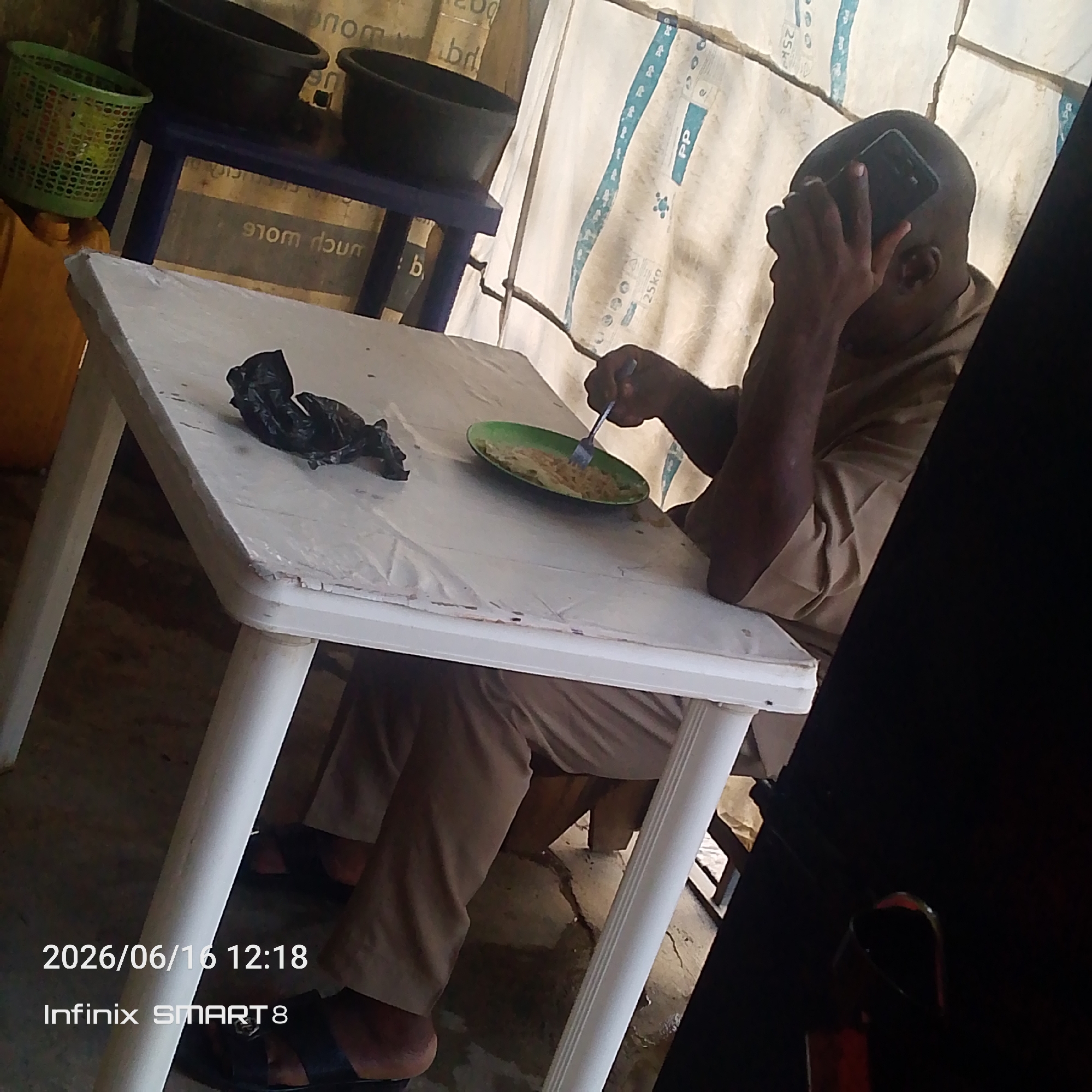 A man sits at a white plastic table with a white plastic tablecloth. He is eating from a green oval plate with a fork. He is holding a black cell phone to his ear with his left hand. The man is wearing a light brown short-sleeved shirt and khaki pants. To the left of the plate is a crumpled black plastic bag. In the background, there is a blue table with two black buckets on it, and a green plastic basket. The backdrop appears to be a sheet of white material with blue markings and text. The scene is outdoors, likely in a casual setting. The image timestamp indicates June 16, 2026, at 12:18 PM, and the device used was an Infinix SMART 8.