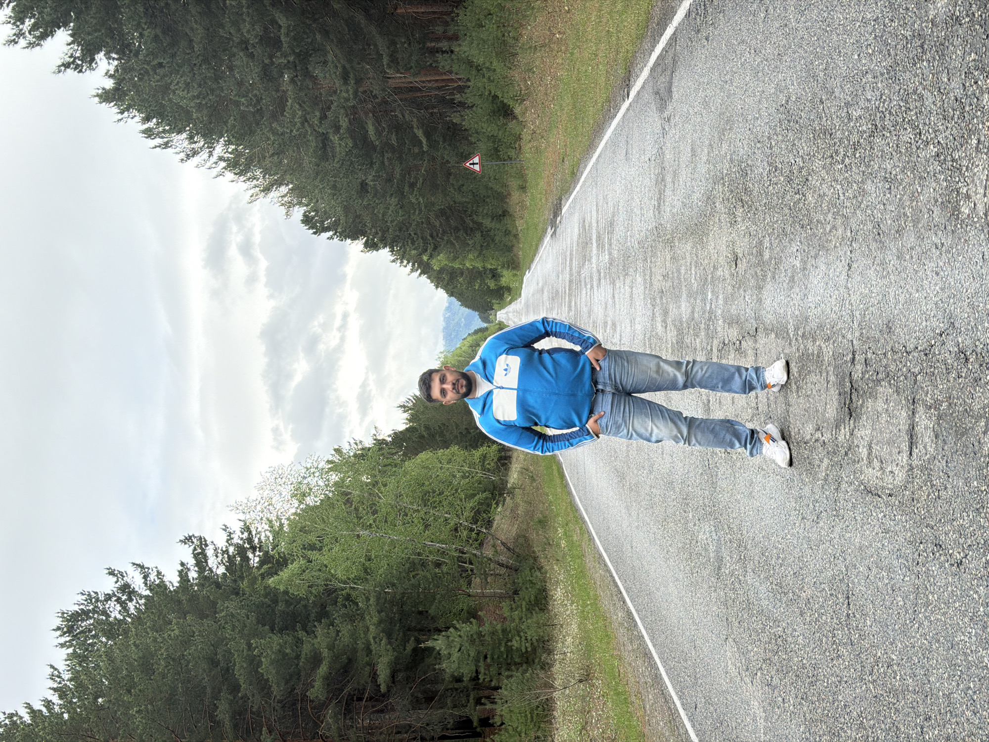 A man is standing on a road in Сизая, Russia. The man is wearing a blue Adidas jacket, jeans, and white sneakers with orange accents. The road is asphalt and has a white line marking the edge. There are trees on both sides of the road. There is a road sign with a cross on it in the distance. The sky is cloudy.