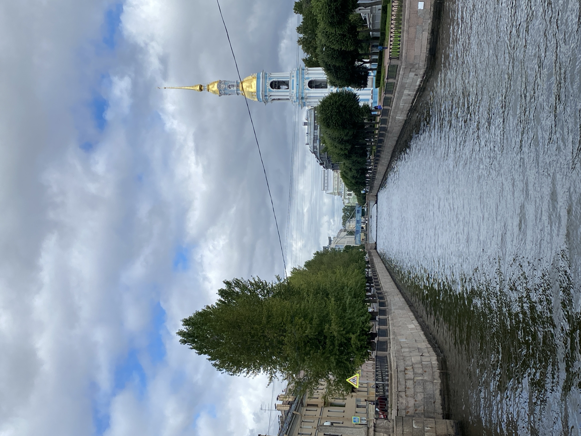 This image captures a scenic canal in Saint Petersburg, Russia, likely the Griboyedov Canal, given its architectural surroundings. The canal, a central feature, reflects the overcast sky above. On the left bank, a row of apartment buildings with classical facades lines the canal, interspersed with tall, leafy green trees. A pedestrian walkway with a black metal railing runs alongside the canal. On the right bank, a prominent landmark, the St. Nicholas Naval Cathedral, stands out with its striking blue and white bell tower topped with a golden spire. A line of trees also borders this side, leading towards the cathedral. A pedestrian path with a manicured green lawn and a railing is visible in front of the trees. A bridge spans the canal in the distance, and power lines crisscross the sky. The overall atmosphere is one of urban tranquility, with a few indistinct figures visible strolling along the pathways.