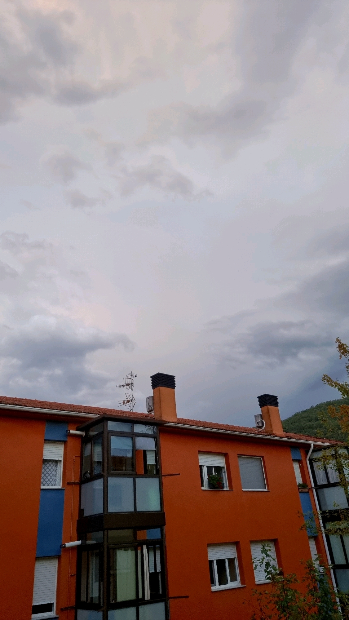 This image shows a section of an orange building with a red tiled roof under a cloudy sky in Sant Joan Les Fonts, Spain. The building features a vertical glass-enclosed structure with multiple windows, as well as several rectangular windows with white shutters on the orange facade. A blue vertical section is visible on the left side of the building, also with white shutters. Two chimneys with black tops rise from the roof, along with a television antenna. The background shows a green, tree-covered hillside. Some autumn foliage is visible on the right side of the building. The scene is devoid of any people or animals.