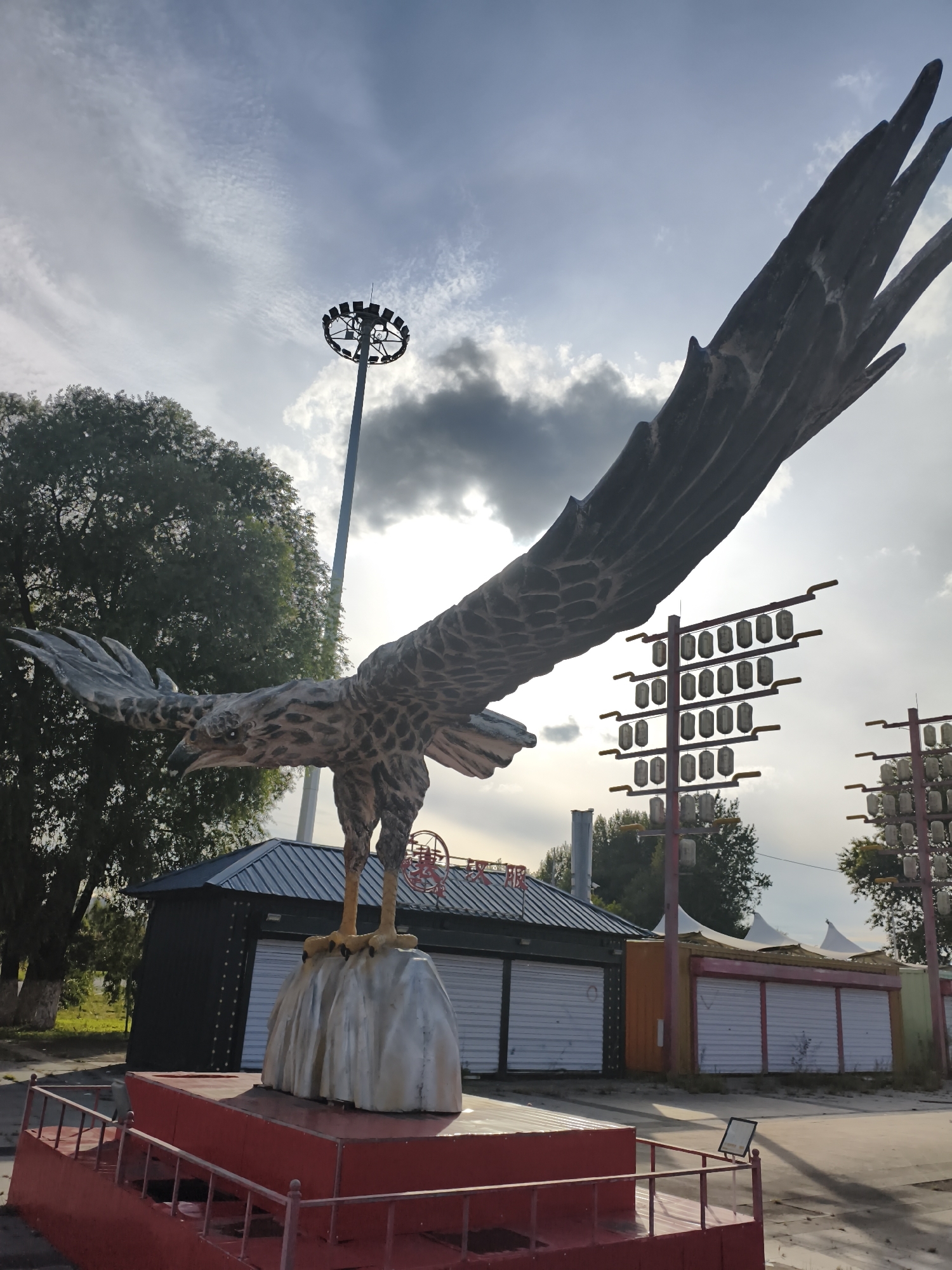 In Yongfeng, China, a large sculpture of an eagle with outstretched wings is the focal point of the image. The eagle stands on a rock, which is mounted on a red platform with railings. Behind the sculpture, a dark building with closed metal shutters is visible, along with several other similar structures. Tall light poles with multiple lamps and string lights are also present in the background. The sky is overcast with scattered clouds. The environment appears to be an outdoor public space, possibly an amusement park or a recreational area that is not currently in operation.