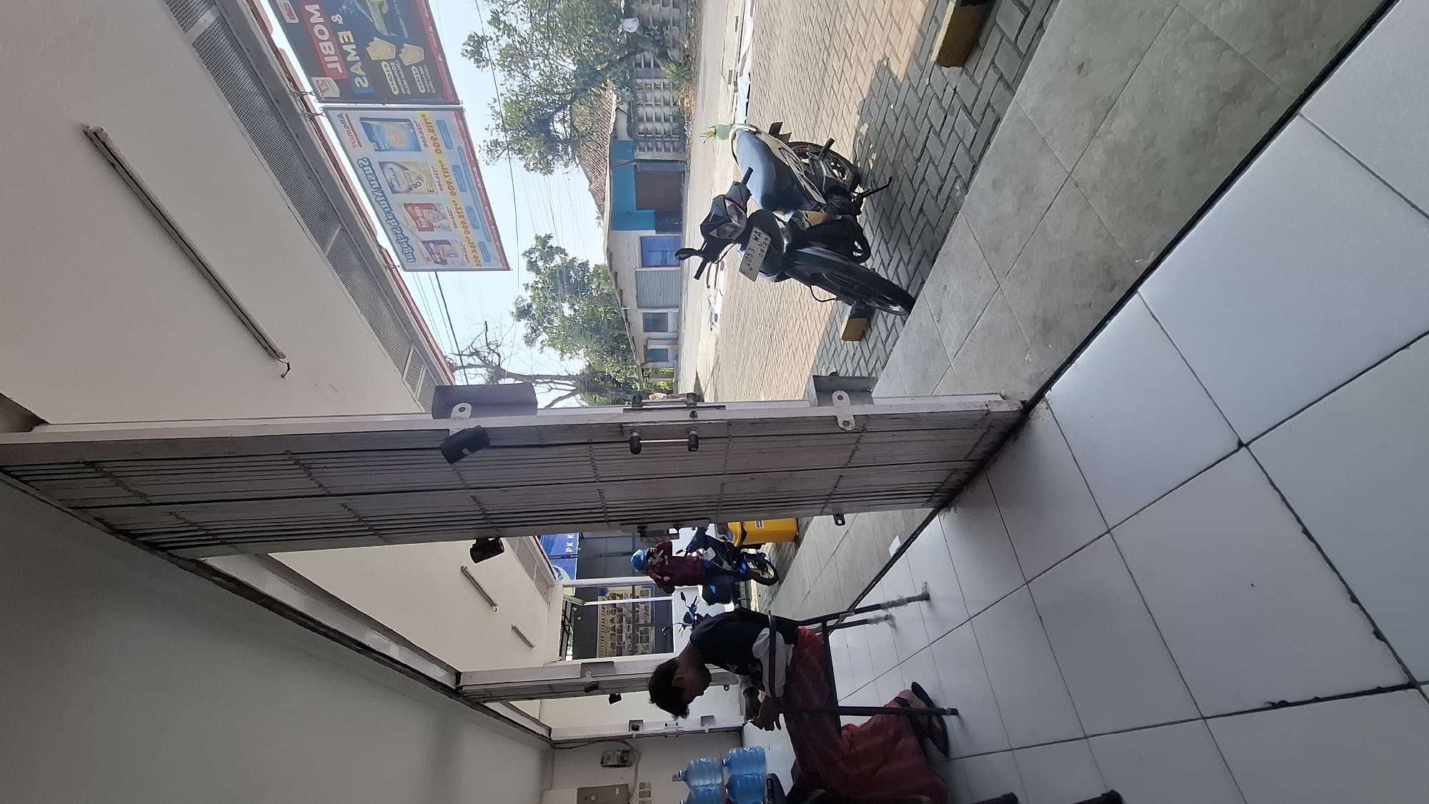 The image captures an outdoor scene in Cieundeur, Indonesia, likely in front of a shop. A man in a dark shirt and red pants is seated on a chair, looking down at his phone or a table in front of him. To his right, a stack of blue water jugs is visible. Further back, another person wearing a red shirt and a blue helmet is standing, possibly engaged with a motorbike. In the foreground, a silver and black motorbike is parked on a tiled walkway. The background shows a street with buildings and trees under a clear sky. The architecture suggests a commercial area.