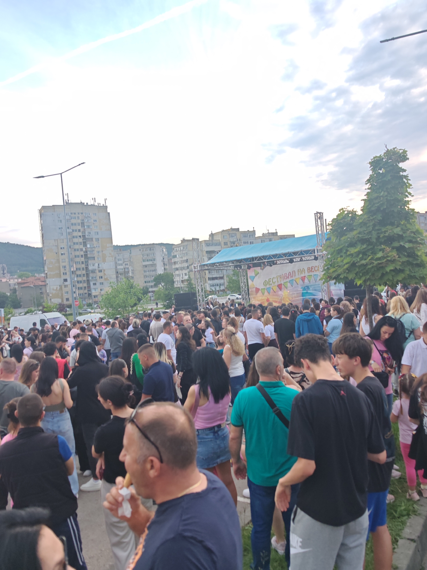 A large crowd of people is gathered in an open space, likely a plaza, in Shumen, Bulgaria. A stage is visible in the background with a banner reading "ФЕСТИВАЛ НА ВЕС", which translates to "Spring Festival". The crowd consists of a diverse group of individuals, including men, women, and children, dressed in casual clothing. Some individuals are looking towards the stage, while others are engaged in conversations or other activities. The buildings are the Communist-era style apartment blocks that define the city skyline. A tree is next to the stage. In the foreground, a man is eating what appears to be some type of food on a stick. The sky is overcast, suggesting it may be late afternoon or early evening.