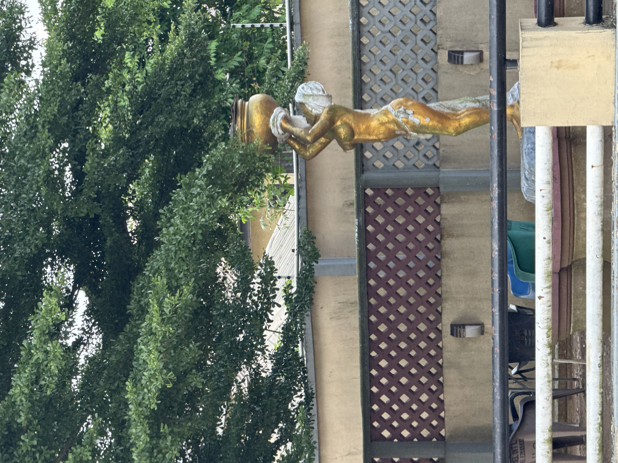 Here's a detailed description of the image:
The image appears to be taken outdoors, likely on a balcony or elevated platform. The predominant feature is a gold-colored statue of a reclining female figure holding a large, ornate pot. The statue is positioned against a backdrop of gray metal lattice and what appears to be a building wall. A tall, dense green tree flanks the left side of the image. The right side includes elements of outdoor furniture, possibly a chair, and other building-related fixtures, such as pipes or supports. The location is stated to be in Ihiagwa, Nigeria, though this cannot be visually confirmed from the image alone.