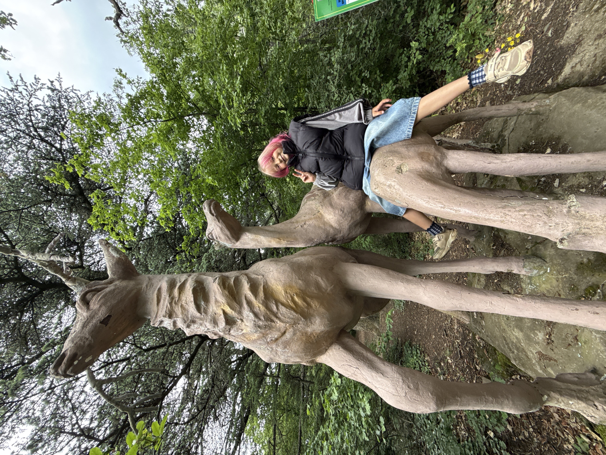 A person with pink hair is riding a statue of a deer in a forest. The person is wearing a black jacket, blue skirt, and patterned socks with tan shoes. The deer statue is a light brown color and appears to be made of stone or concrete. The statue appears to be in a clearing of trees, surrounded by greenery. The location is Unknown, Unknown.