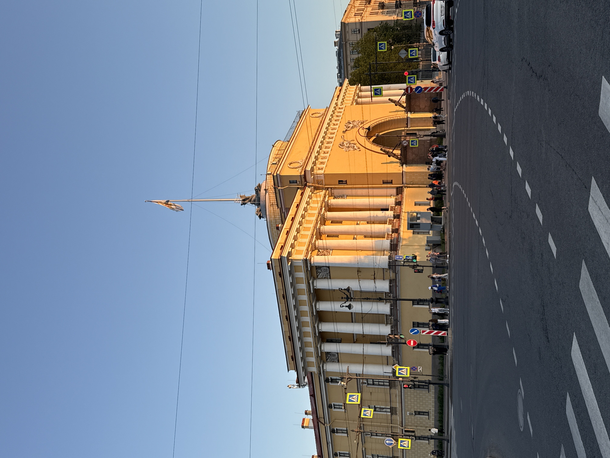 The image depicts a neoclassical building, identified as the Admiralty in Saint Petersburg, Russia, under clear blue sky with direct sunlight. The building features a prominent pale yellow facade with white classical columns. A tall central spire, capped with a ship weathervane, rises above the structure, and a yellow flag, consistent with the Russian Imperial Standard, flies from a flagpole on the spire. The facade is illuminated by a warm, golden light, indicating late afternoon or early evening.
On the right side of the building's visible portion, a large arched entrance with sculptural details, including winged figures blowing trumpets, is prominent. The building's rooflines are visible, featuring red tiles.
In the foreground, a wide asphalt intersection with white dashed lane markings and prominent white pedestrian crosswalks stretches across the frame. Multiple traffic light poles with functional signals (red and green lights visible), streetlights, and various road signs are present. These signs include yellow diamond-shaped pedestrian crossing warnings, a red circular "no entry" sign with a white bar, and a blue circular sign, possibly indicating a mandatory direction. Overhead power lines traverse the upper portion of the sky.
Several groups of pedestrians are gathered on the sidewalks surrounding the intersection, primarily on the far side from the viewer, appearing to wait or converse. They are casually dressed. On the right side of the road, two white vehicles, an SUV and a sedan, are visible, either stopped or parked.