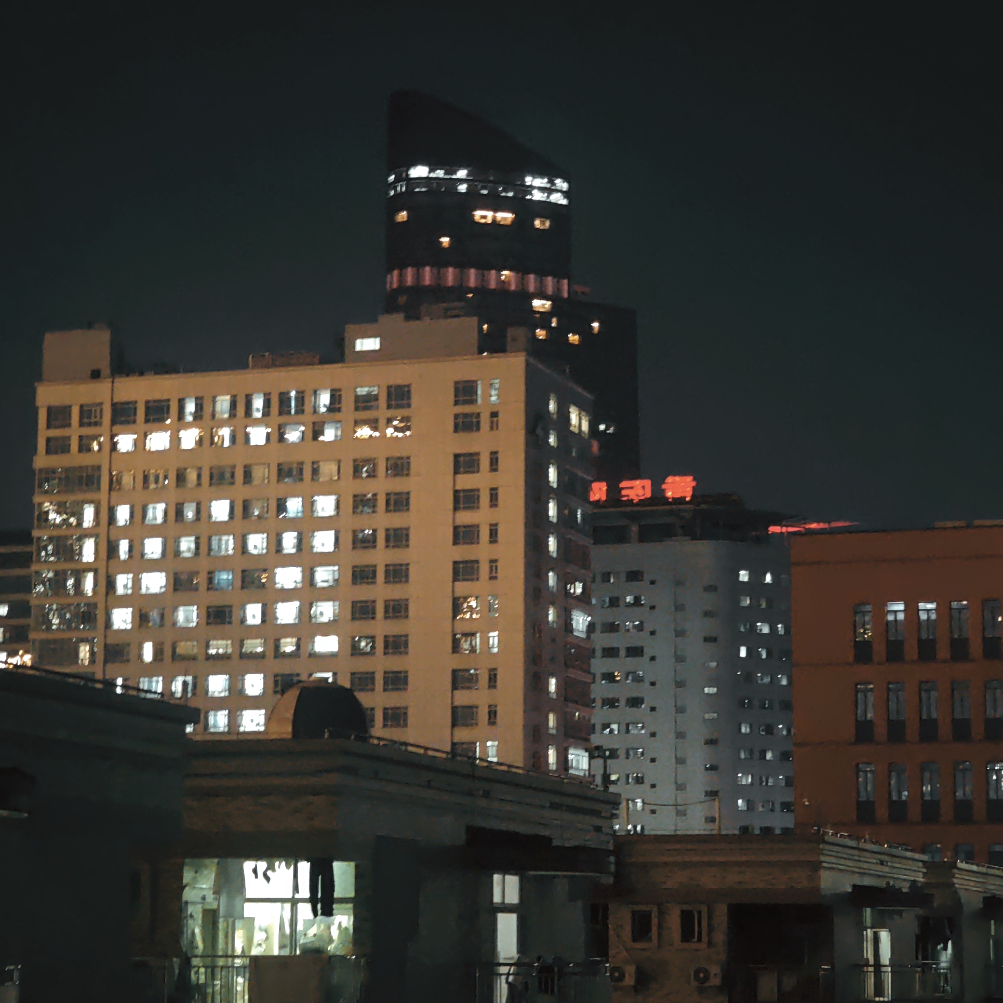 A nighttime urban scene in Yuzhong District, China, showing a view from a lower building towards a cluster of taller buildings. The main building is a large, light-colored apartment complex illuminated from within, with many windows lit. Behind it rises a darker, modern skyscraper with a distinctive top section that has lights. To the right of the apartment complex are other buildings, including one that is light gray with fewer lights and another one in shades of orange. In the foreground, a lower building with a flat roof provides a vantage point, with some laundry hanging on a balcony in the lit window. The night sky is dark.