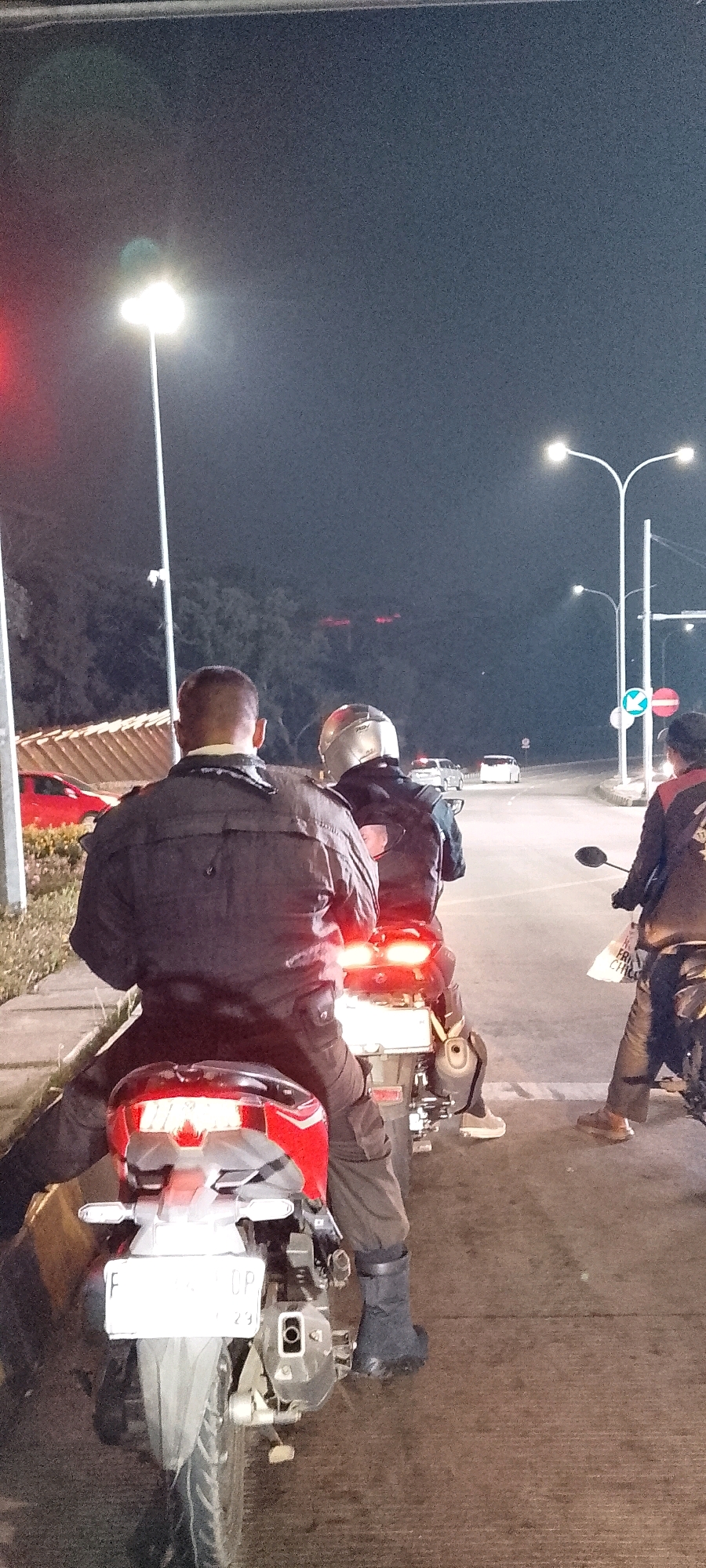In Gunung Putri, Indonesia, at night, three individuals on motorcycles are stopped at a road intersection. The foremost motorcycle, seen from the rear, has a driver wearing dark clothing and a helmet, and is stopped behind a red car. The license plate of this motorcycle is visible, marked with "F . . . DP". The second motorcycle is behind the first, with a driver also wearing dark clothing and a helmet. The third motorcycle is to the right of the first two, with its rider wearing dark pants and a dark jacket, holding a white plastic bag. In the background, streetlights illuminate the intersection, and several cars are visible further down the road. Trees and foliage line the sides of the road, and a red traffic signal is illuminated on the left.