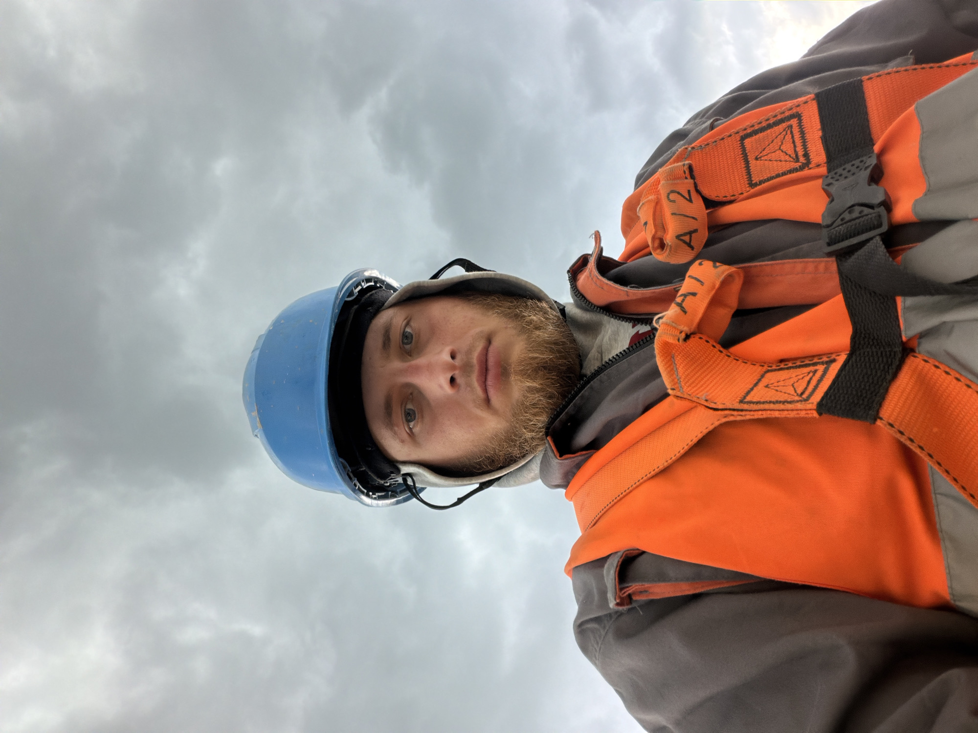 A man wearing a blue hard hat and an orange safety vest is looking down at the camera. He has a beard and is wearing a grey jacket underneath the vest. The background is a cloudy sky. The image was taken in Stróża, Poland.