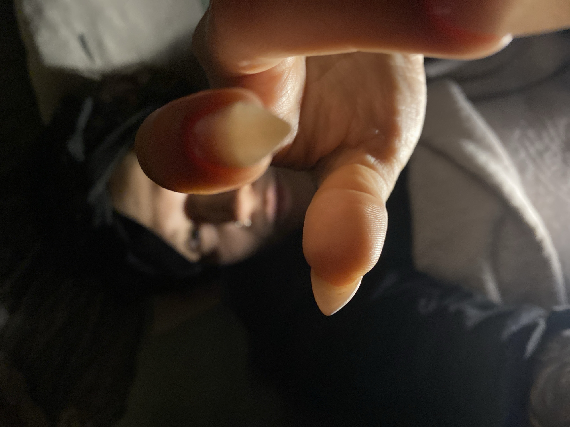 A close-up, distorted perspective shows a person lying down, obscured by fingers extended towards the camera. The person's face is partially visible, wearing what appears to be a dark covering. The background includes a greyish fabric or material. The environment is dimly lit. Location cannot be discerned from the image.