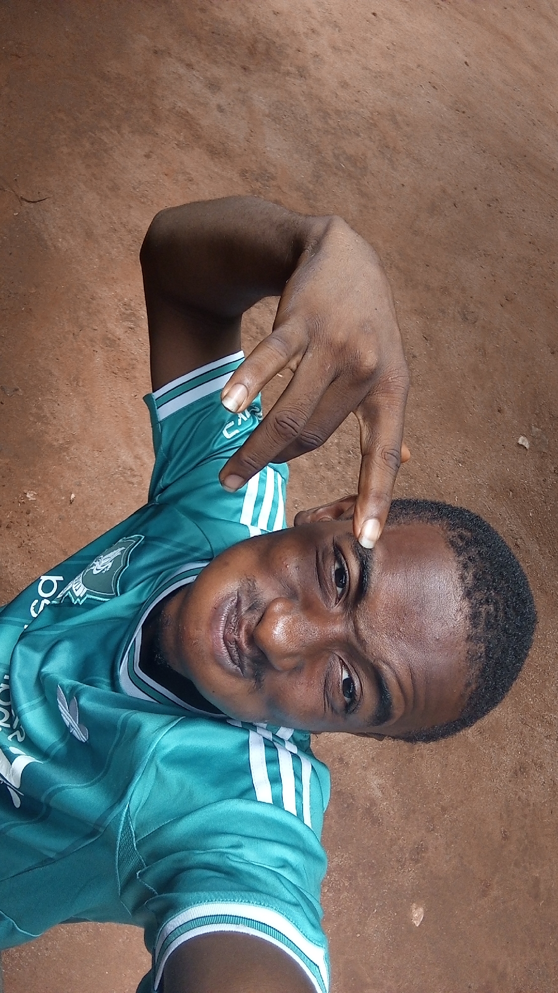 A man with short dark hair and dark skin is looking directly at the camera with a slight smile. He is wearing a green and white Adidas sports shirt. His right hand is raised, with his index finger touching his forehead. The background appears to be a reddish-brown dirt surface. The location is Unknown, Unknown.