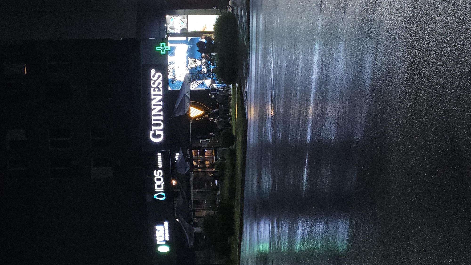 The image depicts a street in Plovdiv, Bulgaria at night. It's raining and the pavement is wet, reflecting the lights of the buildings. On the left side of the street are buildings with illuminated signs, including a pharmacy with a green cross, a "Guinness" sign, an "IQOS" sign, and another sign, partly obscured, in green. There are also some outdoor tables and chairs visible under umbrellas. The lighting is a mix of bright artificial lights from the signs and the muted reflections on the wet ground. The general atmosphere suggests a commercial area or a street with businesses and restaurants.