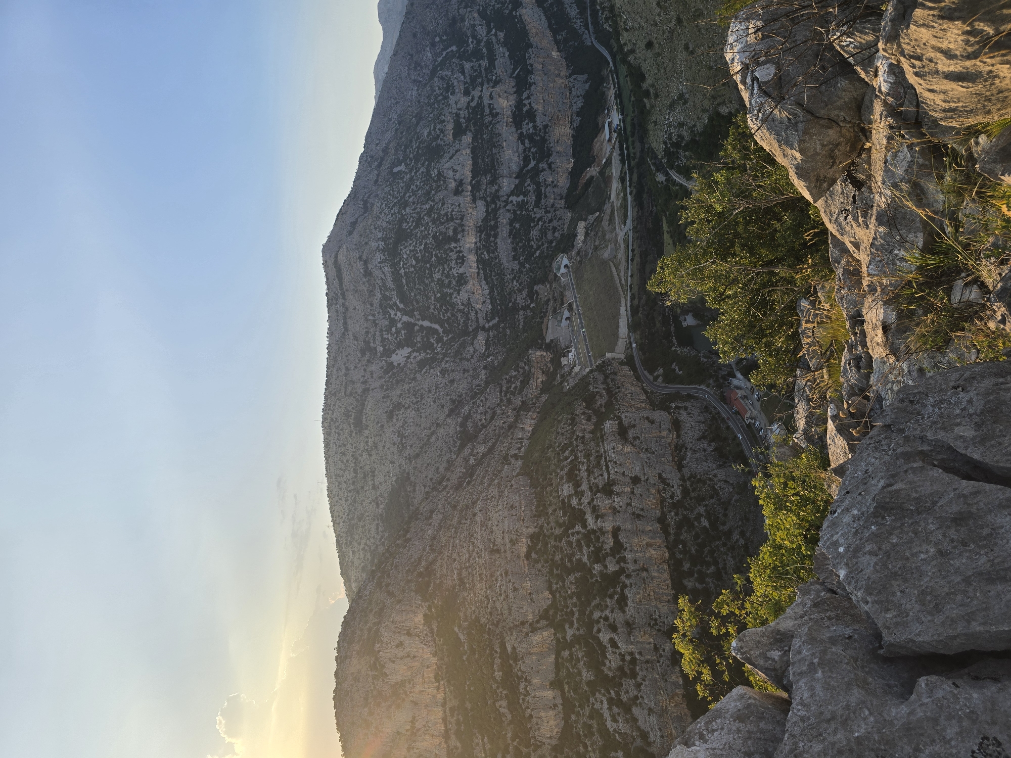 The image captures a panoramic view of a rugged mountain landscape with a winding road and a dam. The perspective is from a high vantage point, looking down into a valley. The mountains are characterized by layered rock formations and sparse vegetation, with some trees and shrubs dotting the slopes. The sun is setting or rising, casting a warm glow and long shadows across the scene, with rays of light filtering through clouds.
In the valley below, a road snakes its way through the terrain, with a few cars visible. A dam structure is prominent, with tunnels carved into the mountainside adjacent to it. A small building complex appears near the dam. The foreground is dominated by large, weathered rocks and dry grasses, suggesting a natural, mountainous environment.
While the image depicts a scenic landscape, it does not provide specific visual cues that definitively place it within the city of Podgorica, Montenegro. The scene is more characteristic of a natural, mountainous region.