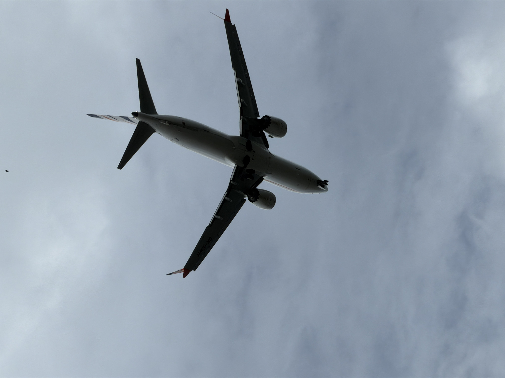 An airplane is captured from a low-angle perspective, silhouetted against a cloudy sky. The aircraft appears to be in mid-flight, with its underside and wings prominently visible. The sun is obscured by the dense cloud cover, creating a diffused light that casts the airplane in shadow. The weather appears overcast, contributing to the muted tones of the scene.
The airplane's design is typical of a commercial jet, featuring two engines mounted under its wings and a distinct tail fin. On the wingtip, the registration "OK-SWH" is visible. The overall impression is one of a vast, empty sky with the sole, dominant presence of the flying machine. The scene is devoid of any people or ground-level context, focusing entirely on the aerial perspective. The presence of an airplane in flight in this location suggests normal air traffic. The time of day cannot be precisely determined due to the overcast conditions.