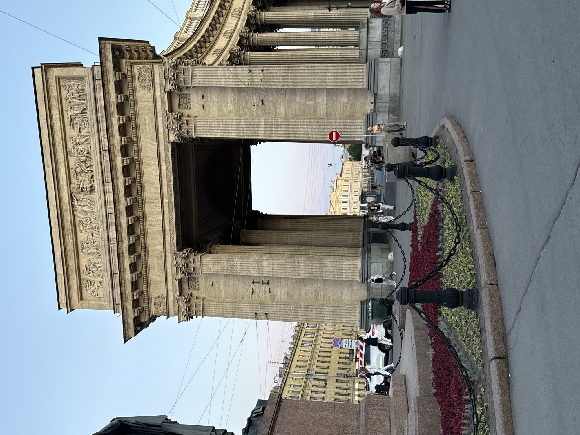 The image depicts a street scene in Saint Petersburg, Russia, with the Kazan Cathedral's colonnade prominently featured. The perspective is a low-angle shot, looking up towards the monumental architecture. The colonnade, made of stone, is adorned with intricate carvings and classical columns. Sunlight casts shadows, suggesting it is either morning or late afternoon.
Several people are visible in the scene, some walking on the paved area and others standing. They appear to be tourists or locals engaged in everyday activities. A white car with red and blue stripes is parked on the street, and a red and white circular sign with a horizontal bar (a "no entry" or "do not enter" sign) is mounted on a pole near the colonnade.
In the foreground, a landscaped area with red and green plants is separated by black metal fencing with chains. The overall atmosphere is one of historical grandeur combined with urban life. The sky is clear and light blue, indicating fair weather. The architecture and the presence of people suggest a public space where visitors can admire the city's landmarks.