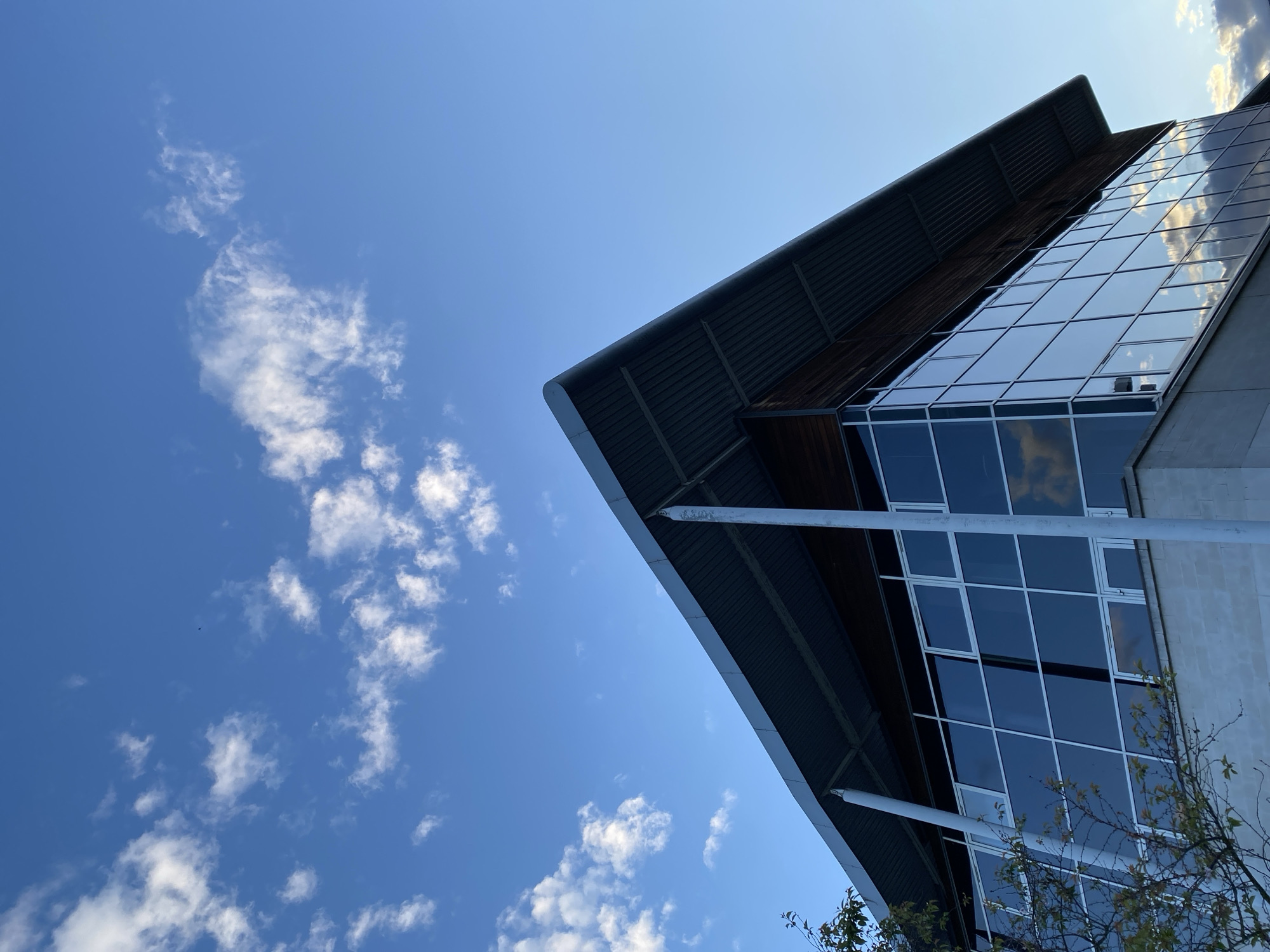 The image depicts a modern building with a sharp, angular roof and a facade largely composed of glass windows, reflecting the blue sky and clouds. The building's architecture is characterized by clean lines and a minimalist design. The structure appears to be positioned at an angle, with the camera looking upwards towards it against a clear blue sky dotted with small, scattered cumulus clouds. The scene lacks any visible human presence. The location of the scene is indeterminable, as the building in the image can not be definitively placed in London, or any other city, without further details.