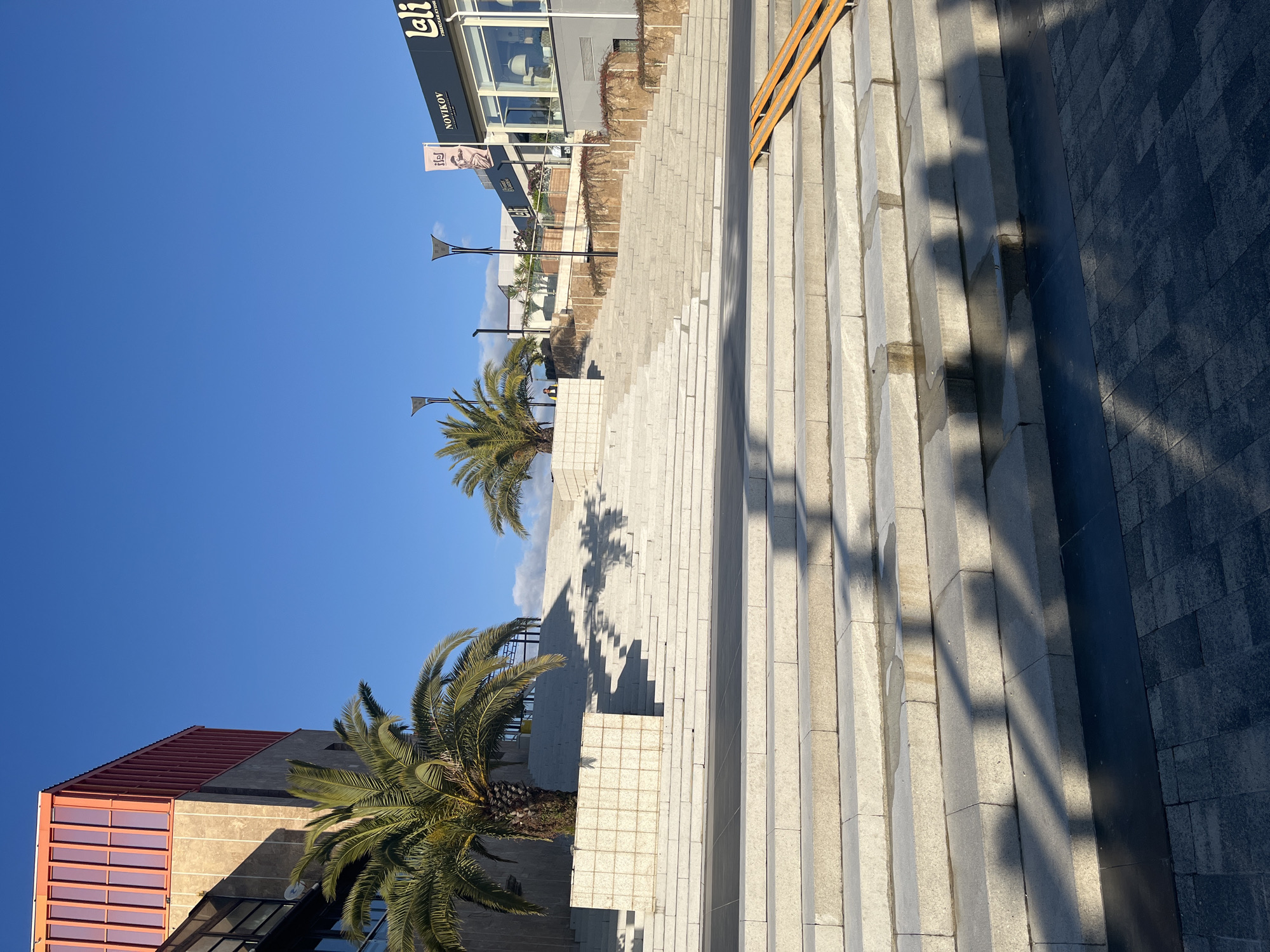 The image depicts an outdoor scene in what appears to be a modern urban area. The scene is set in Sochi, Russia based on visible storefront signage. There are steps leading uphill from a plaza made of dark grey square pavers. Palm trees are located along the stairway and adjacent to the steps. Overhead are shadows cast onto the steps from unseen objects, with a building visible in the background on the left. A sign for "NOYIKOY" is visible above the steps. The sky is blue, indicating a sunny day. The image seems to be taken at ground level, slightly angled upwards.