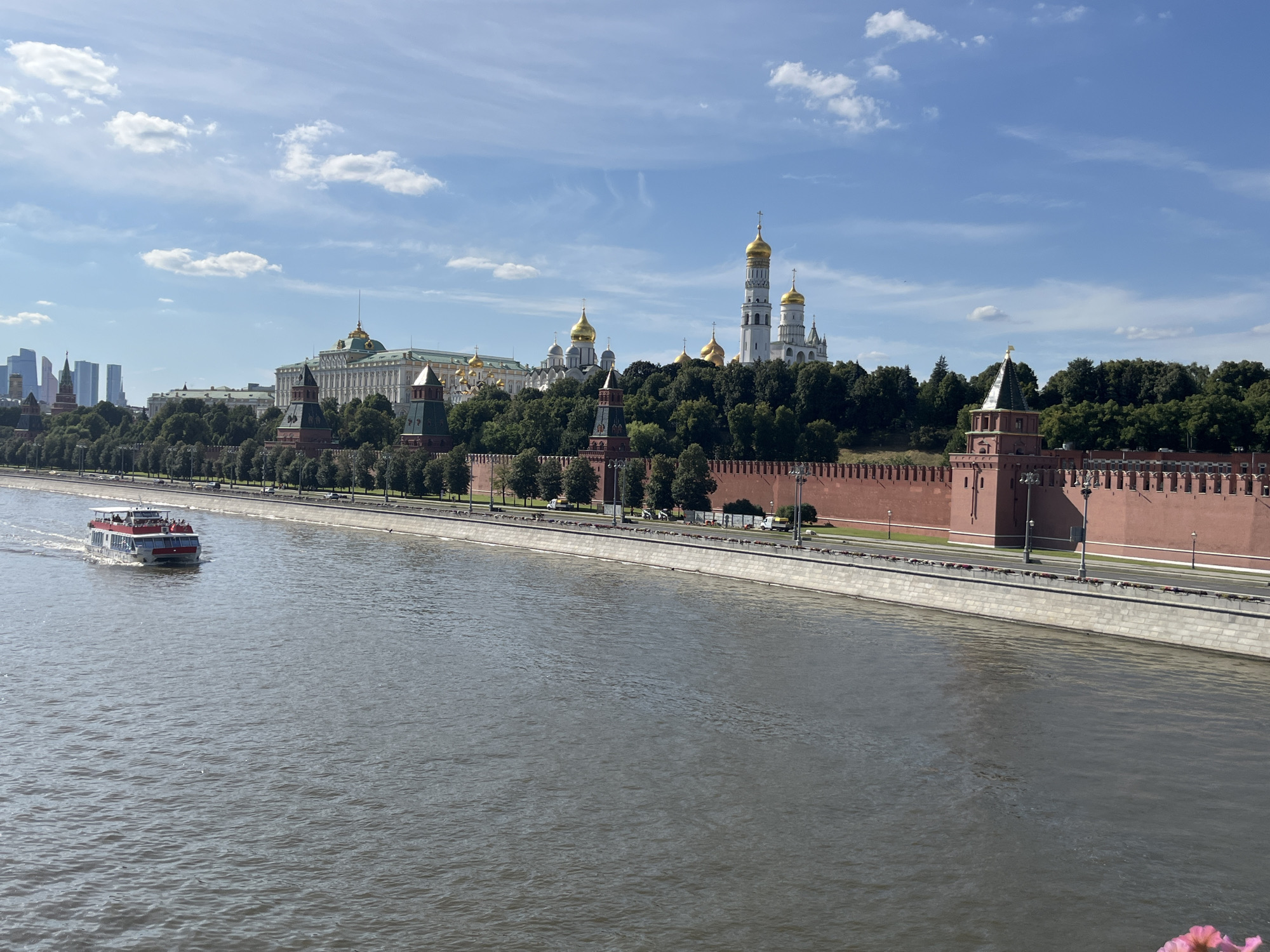 A wide, grey river flows in the foreground, with ripples on its surface. A white and red passenger boat with multiple decks is moving left to right across the river, leaving a white wake. In the midground, a long, red brick wall with crenellations and numerous towers stretches along the riverbank. Behind the wall, a dense line of green trees partially obscures various buildings, including some with green roofs and domes, and several prominent white and gold-domed Orthodox churches. Further in the background, modern skyscrapers are visible on the left side of the frame under a blue sky with scattered white clouds. The scene appears to be a cityscape, likely Moscow, Russia, given the distinctive architecture of the Kremlin and its surrounding structures.