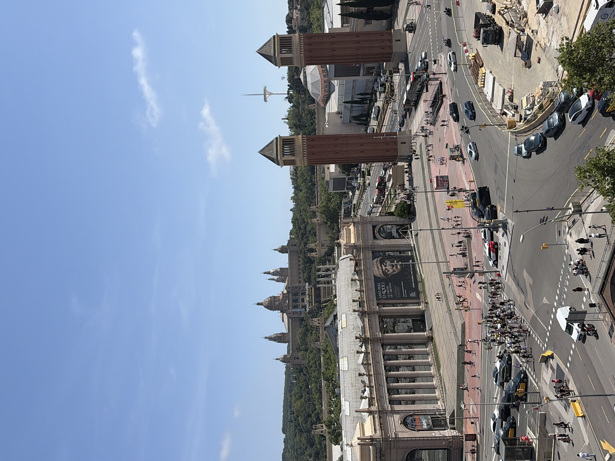 The image captures a high-angle view of Barcelona, Spain, displaying its urban landscape. The foreground features architectural structures with columns, likely part of a museum or grand building, adjacent to a wide pedestrian promenade. A large poster hangs on the building’s facade. Beyond this lies a tree-lined area leading to the Museu Nacional d'Art de Catalunya (MNAC), a prominent building with multiple domes. Further in the background stand the Venetian Towers, brick-built structures that flank a square. In the distance, the communications tower Torre Telefonica, is visible. The road is busy with both pedestrian and vehicular traffic. Construction activity is noticeable in a section of the street. The sky is clear blue with scattered clouds.