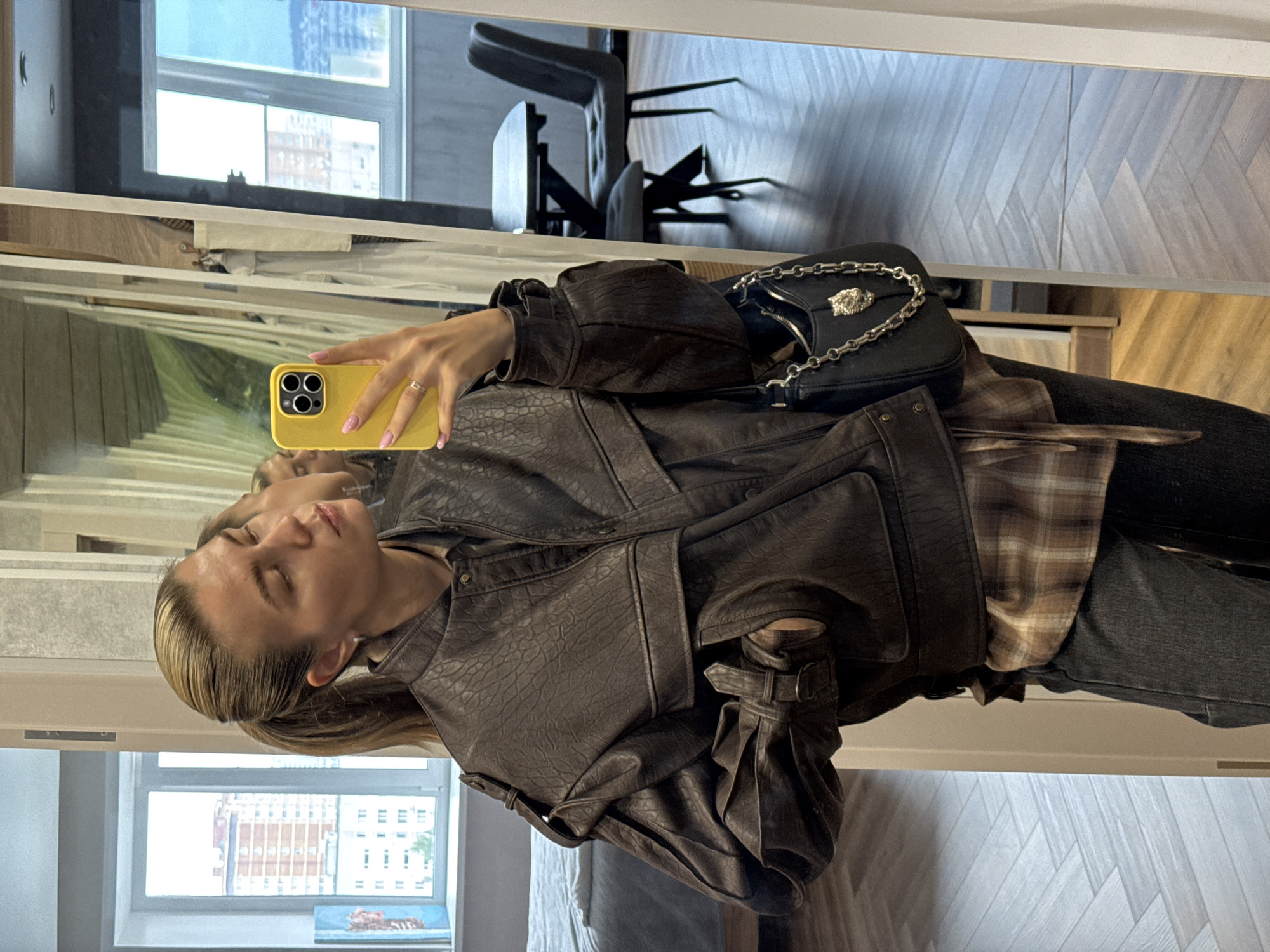The image is a vertical, slightly angled, full-body mirror selfie of a young woman. She is wearing a dark brown faux leather jacket with an embossed pattern, a plaid shirt underneath, and dark wash jeans. She is holding a bright yellow iPhone in her right hand, which is angled towards the camera. The phone has a case with a prominent camera module. A black handbag with a silver chain strap and a lion's head embellishment is slung over her left shoulder.
The woman has blonde hair pulled back, and her eyes are closed, giving her a relaxed or contemplative expression. She is wearing a ring on her left ring finger. The background, as seen in the mirror's reflection, appears to be an indoor setting with a modern aesthetic. Visible elements include a window with a view of city buildings, wooden flooring with a herringbone pattern, and furniture such as chairs and a table, suggesting a residential or studio environment. The lighting appears to be natural, coming from the window, and it's difficult to determine the time of day or weather conditions. There is no visible text in the image.