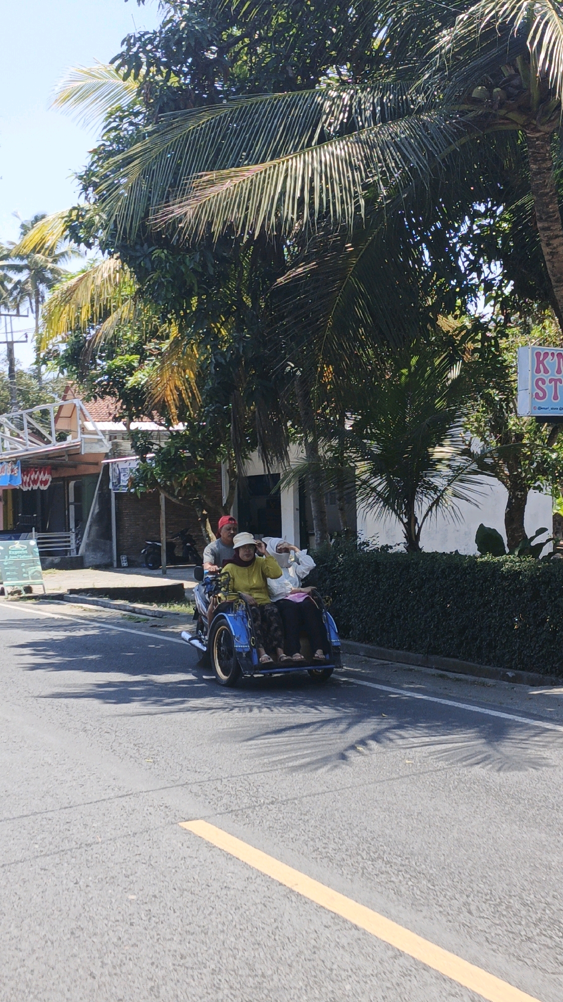 In Desa Wonoharjo, Indonesia, a three-wheeled motorcycle, likely a bajaj or tricycle, navigates a paved road on a sunny day. The vehicle is blue with a single wheel at the front and two at the rear, forming a passenger compartment. Three individuals are seated on the vehicle. The driver, a man wearing a red cap, is at the handlebars. Behind him, two women are seated in the passenger area. The woman in front is wearing a yellow long-sleeved shirt and a light-colored hat, with her left hand raised to adjust her hat. The woman behind her is partially obscured. The vehicle and its occupants are casting shadows on the road. Lush green foliage, including palm trees, lines the right side of the road, obscuring buildings. On the left side of the road, there are buildings with red roofs and awnings, suggesting a commercial or residential area. A white sign with pink lettering is visible on the right side of the road, partially obscured by trees, reading "KT... ST...". The road surface is asphalt, marked with a yellow line. The sky is clear and blue.