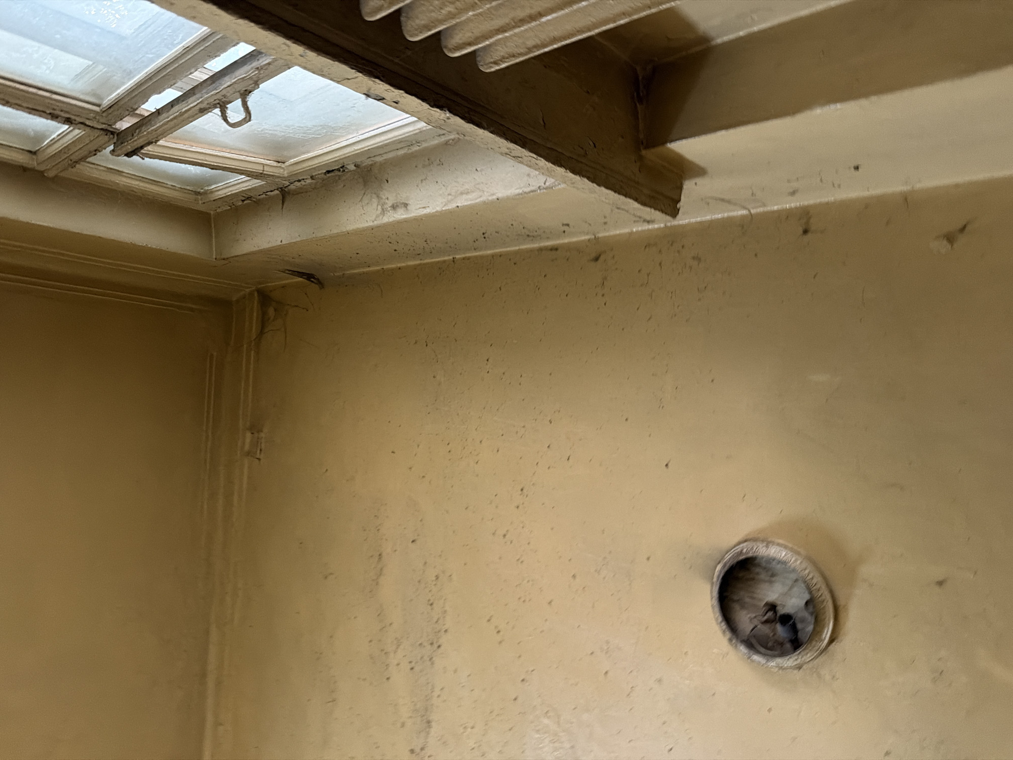 This image depicts the interior of a dimly lit, aged room, likely in an apartment building in Moscow, Russia, showing signs of disrepair. A light yellow-brown wall dominates the frame, marked by numerous dark specks and streaks, suggesting accumulated dirt and dust. In the lower left quadrant, a circular, recessed fixture, possibly an old electrical outlet or speaker grille, is visible, its interior dark and somewhat obscured. The upper right portion of the image features an old, wooden-framed window, partially open, revealing a hazy, light-colored view of the exterior. The window frame itself appears weathered and peeling. A narrow radiator or vent is positioned beneath the window on the right side. The ceiling, visible in the upper left, is also painted a similar light yellow-brown, with a decorative molding along the wall junction that shows cracks and peeling paint. Cobwebs are faintly discernible in the upper corner near the window. The overall impression is one of neglect and the passage of time in an urban dwelling.