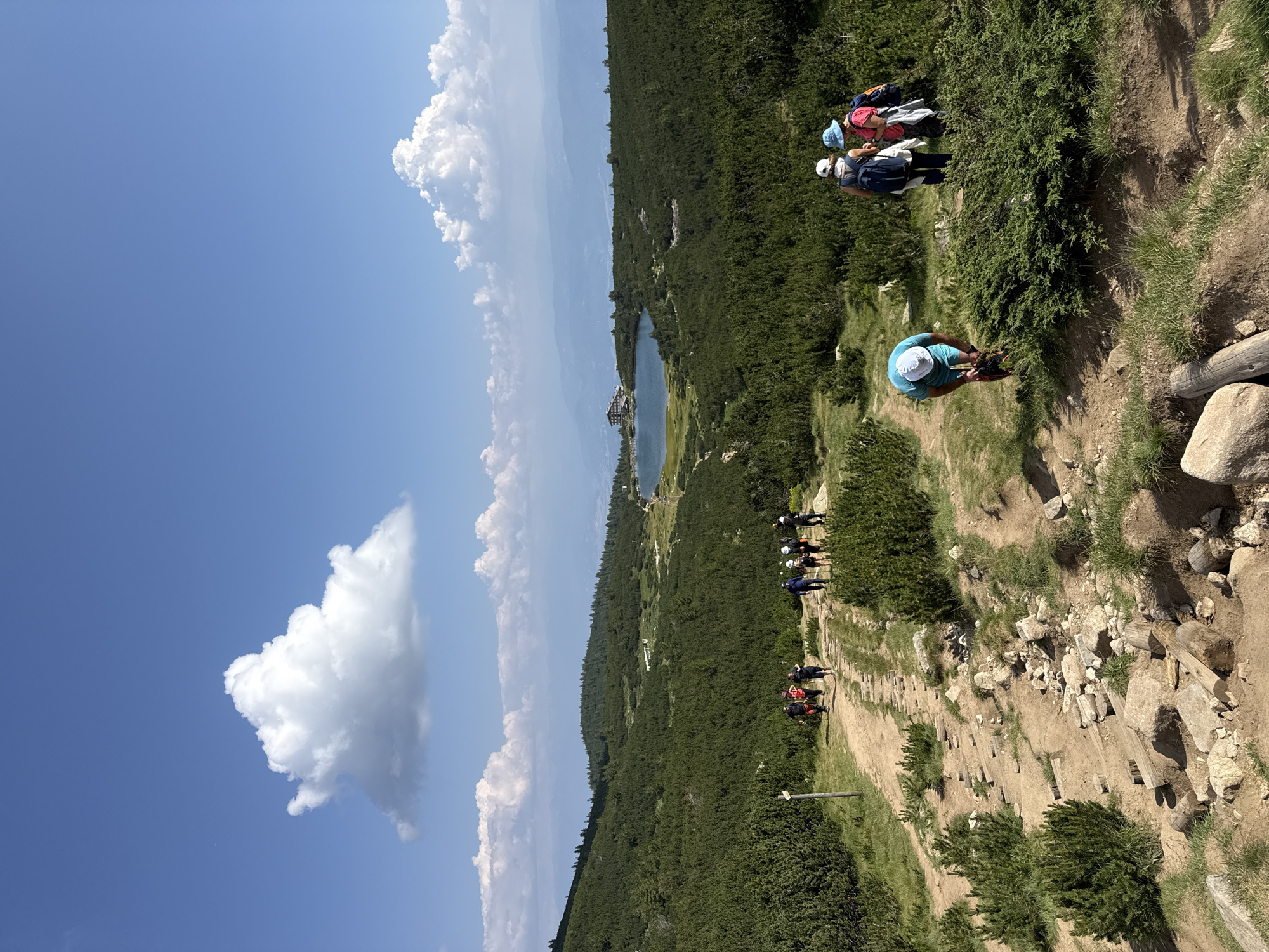 A group of hikers are walking on a dirt path in a mountainous area. The path is rocky and has some wooden steps. The area is covered in dense green bushes and trees. In the distance, there is a lake with a building on its shore, possibly a lodge or hotel. The sky is blue with scattered white clouds. One hiker in the foreground is bending down to adjust their bag, while others are walking further up the path. The scene appears to be in the Pirin Mountains near Bansko, Bulgaria, a popular hiking destination.