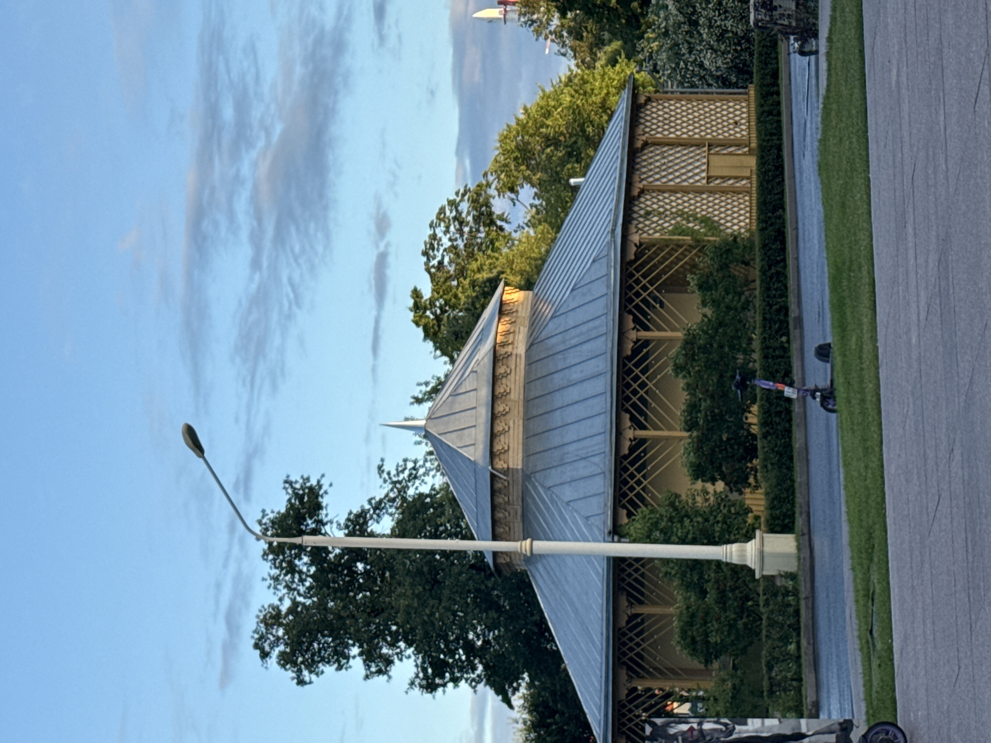 The image depicts a circular pavilion with a grey metal roof and ornate wooden latticework on its sides, set amidst lush green trees. A tall white lamppost stands prominently in front of the pavilion. In the foreground, a paved area transitions to a grassy lawn, with a road bordering the pavilion. Two electric scooters are visible on the road, and a portion of a large mural or advertisement is present on the left edge of the frame. The sky above is a soft blue with scattered clouds, suggesting it might be early morning or late afternoon. This pavilion is located in Gorky Park, Moscow, Russia.